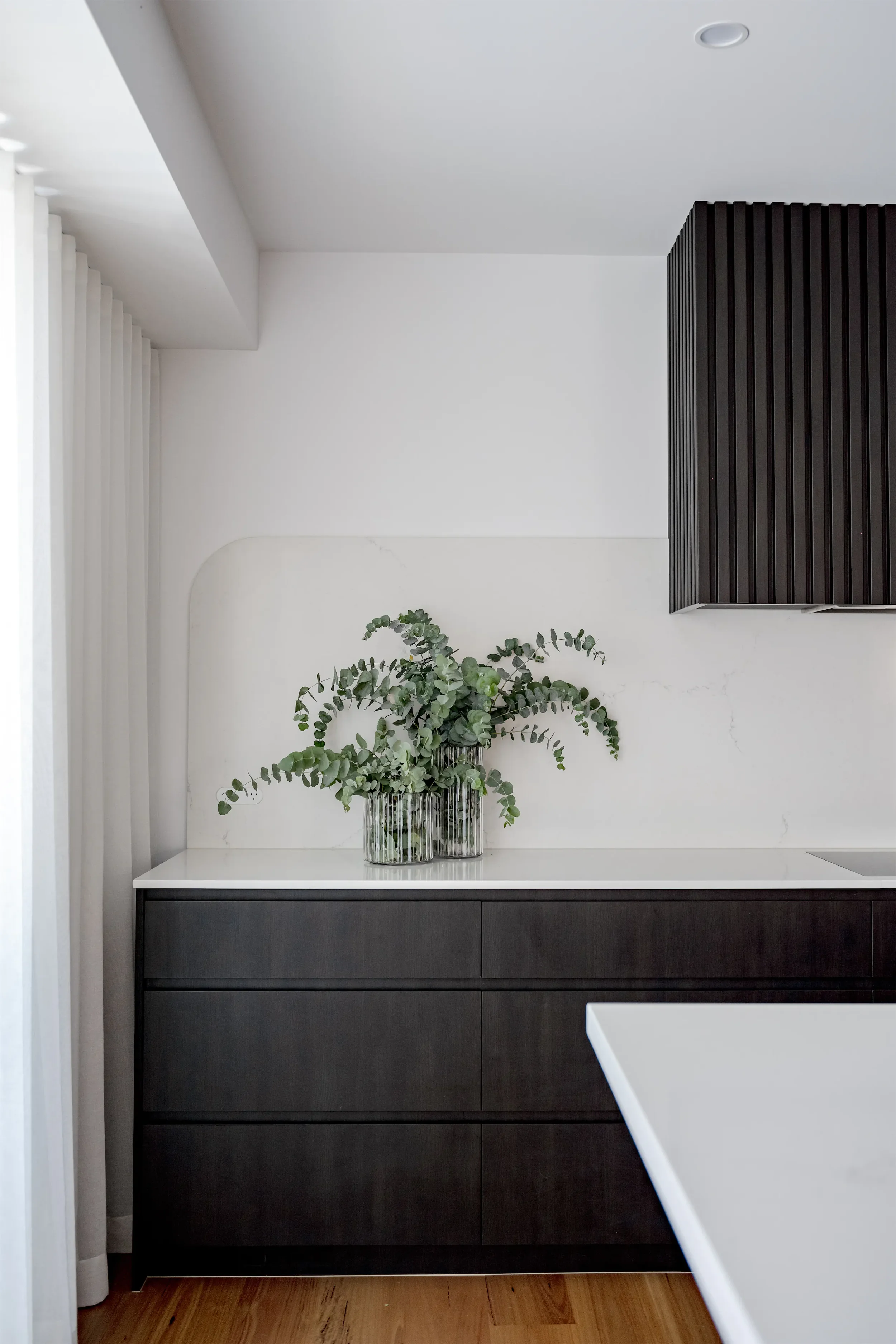 Minimalist kitchen with white marble countertop, black cabinetry, and a clear glass vase with green eucalyptus leaves, against white walls and a light wood floor.