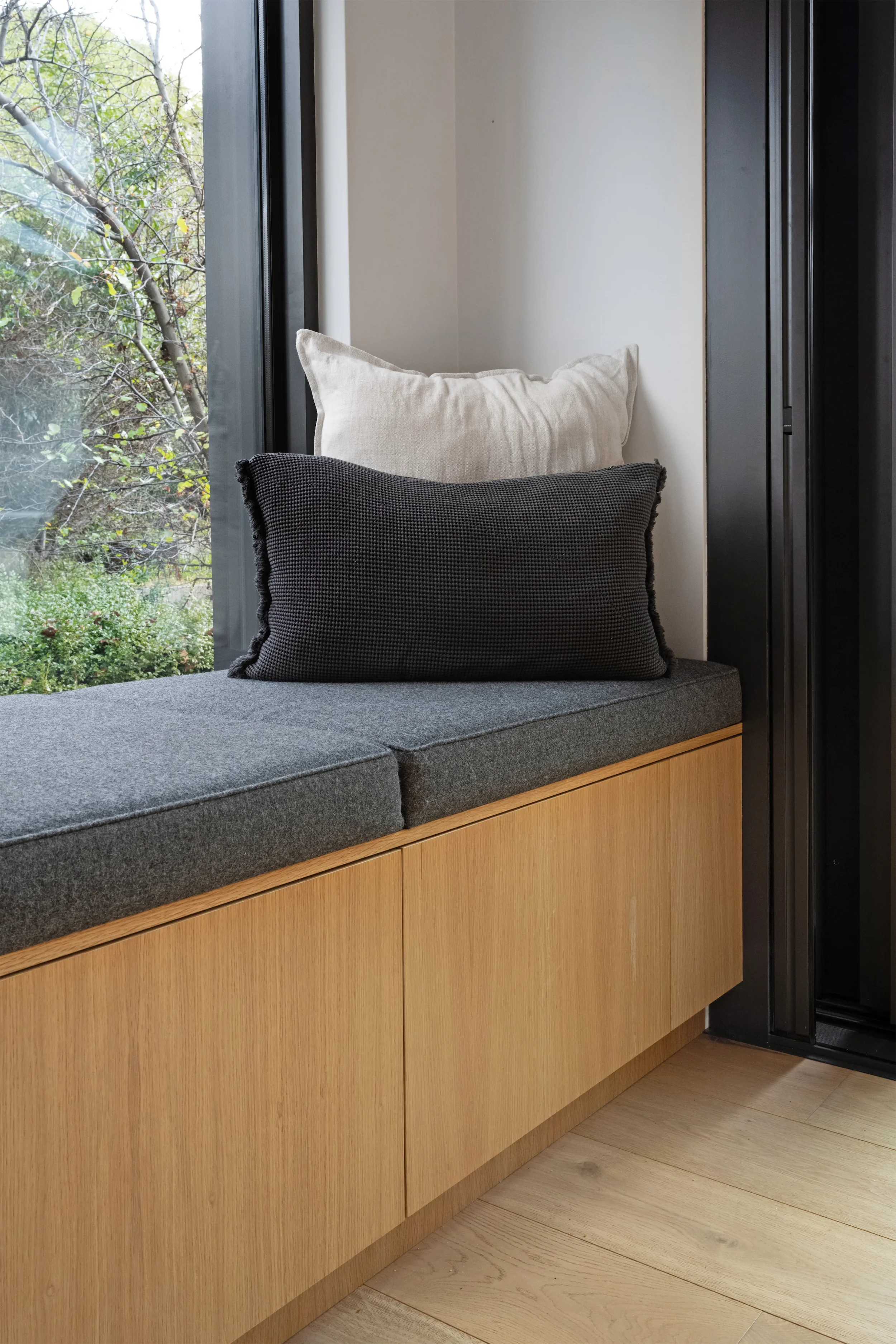 Close-up of a window seat with a dark gray cushion and two pillows, one beige and one black, by a window with a view of trees and greenery outside, and a wooden cabinet below the seat.