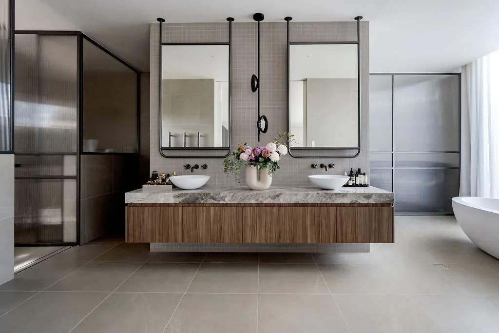 Modern bathroom with a double vanity featuring a marble countertop, two vessel sinks, and two large rectangular mirrors. A vase with pink and white flowers sits between the sinks. The bathroom has beige tiled floors, frosted glass shower enclosures, and a freestanding bathtub on the right side.