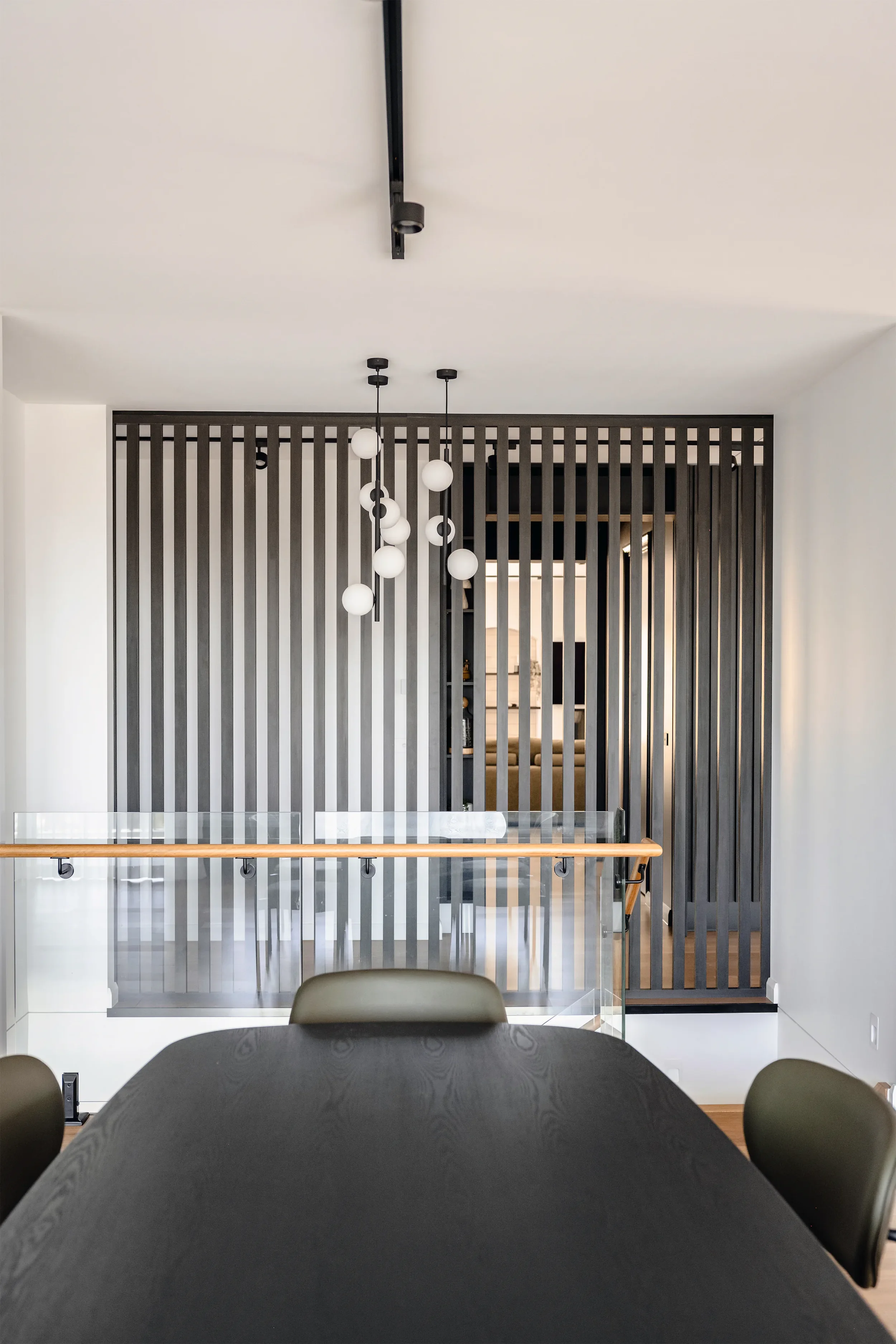 Interior view of a modern dining room with a dark table, green chairs, a glass railing, a wall with vertical dark wood slats, pendant lights, and a doorway leading to another room with a staircase.