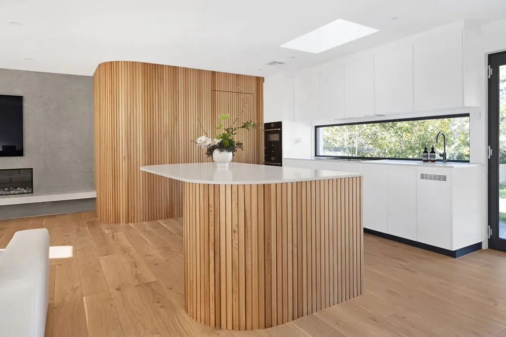 Modern kitchen with white cabinets, black trim, wooden island with a white countertop, large window with greenery outside, and a wood-paneled curved wall.