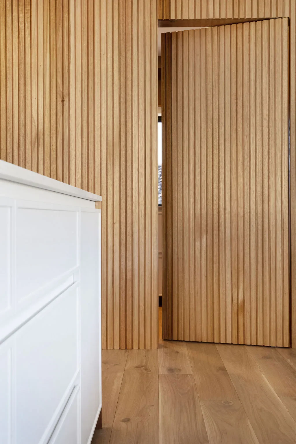 Wooden door with vertical slats slightly open, revealing part of a window with curtains in the background.