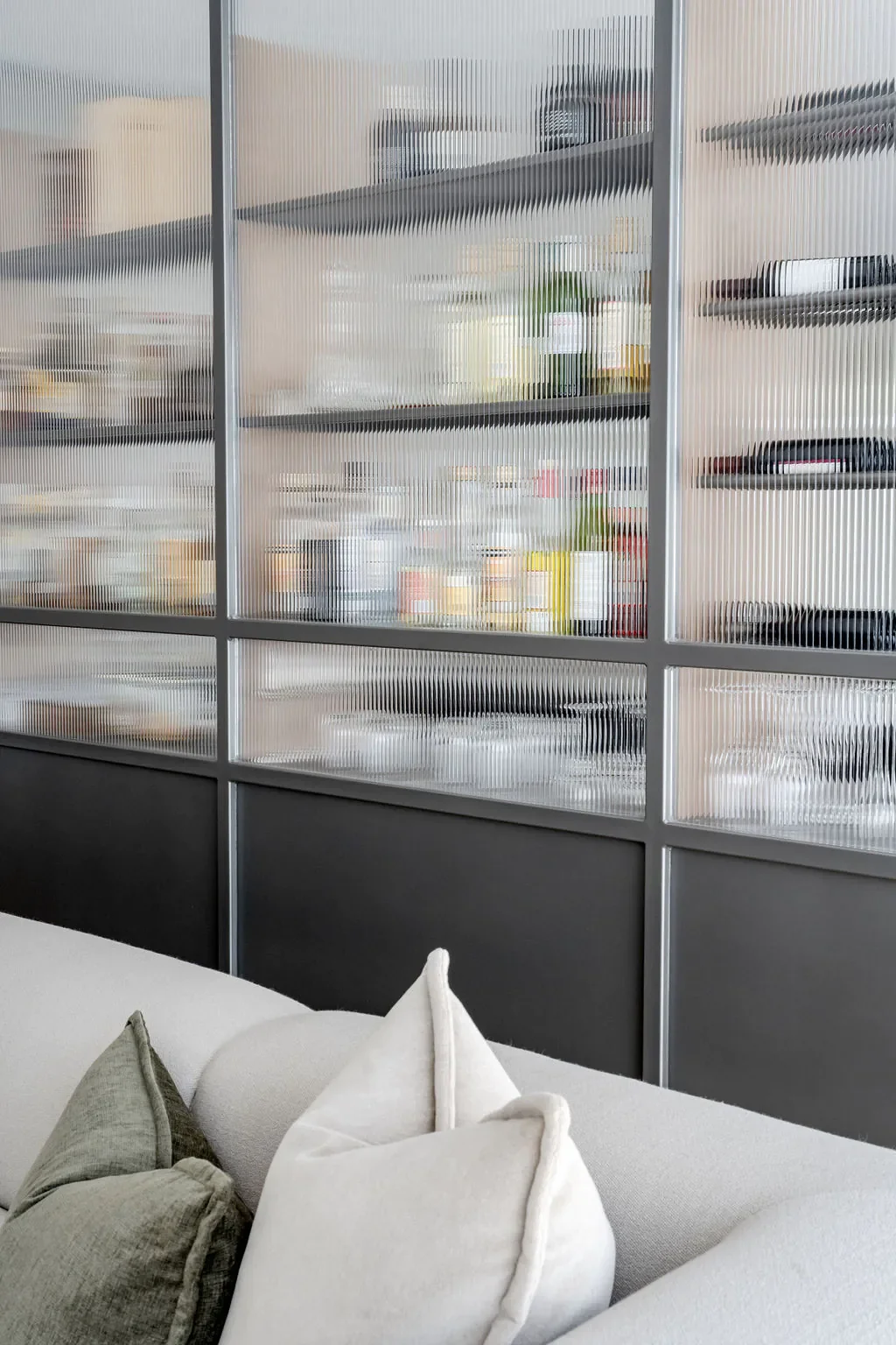 Interior view showing frosted glass shelves with books, plants, and decorative items behind a partition wall, with a white sofa and pillows in the foreground.