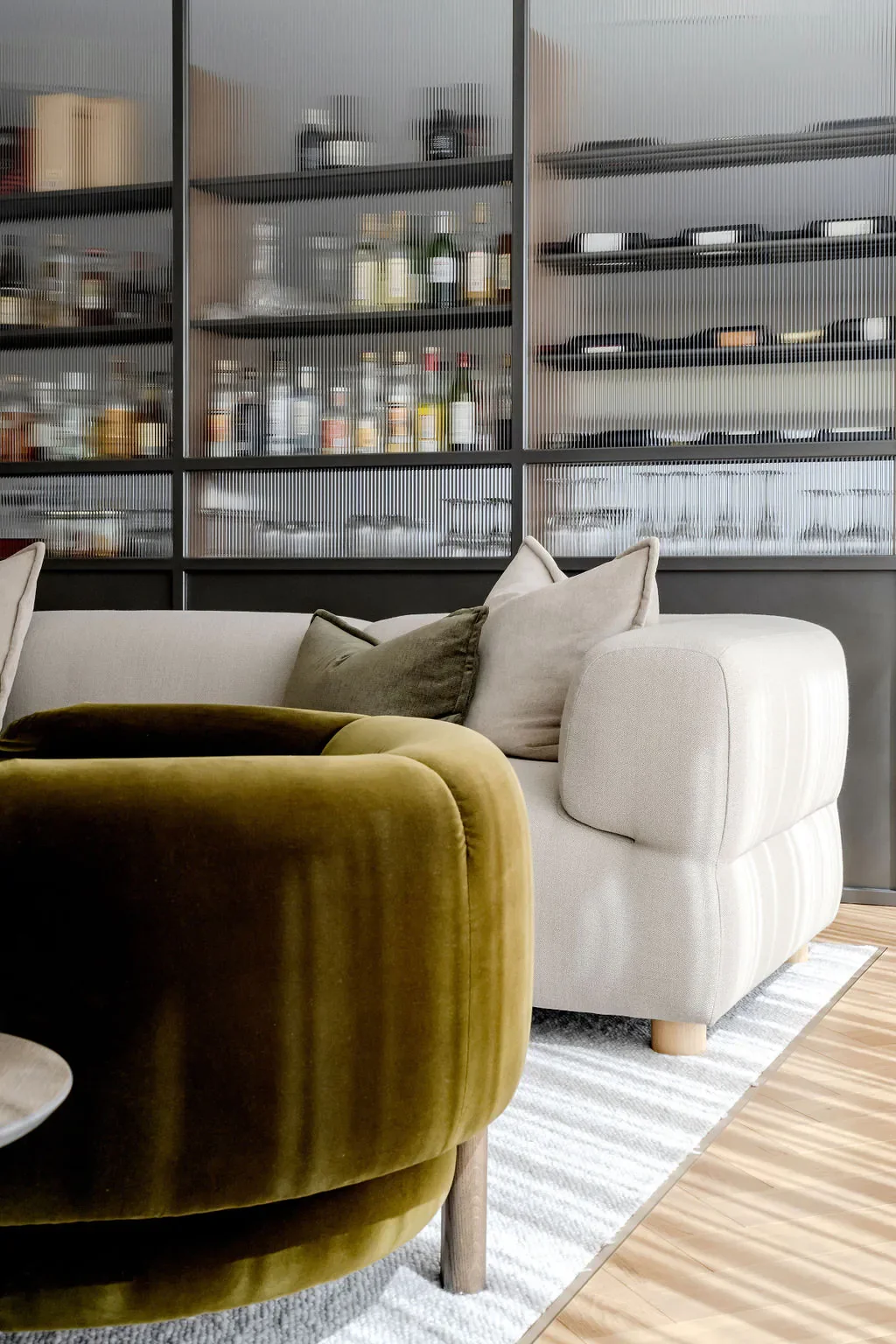 A cozy living room with a white sofa and an olive-green velvet armchair on a striped rug, with a glass-fronted cabinet in the background containing bottles and dishes.