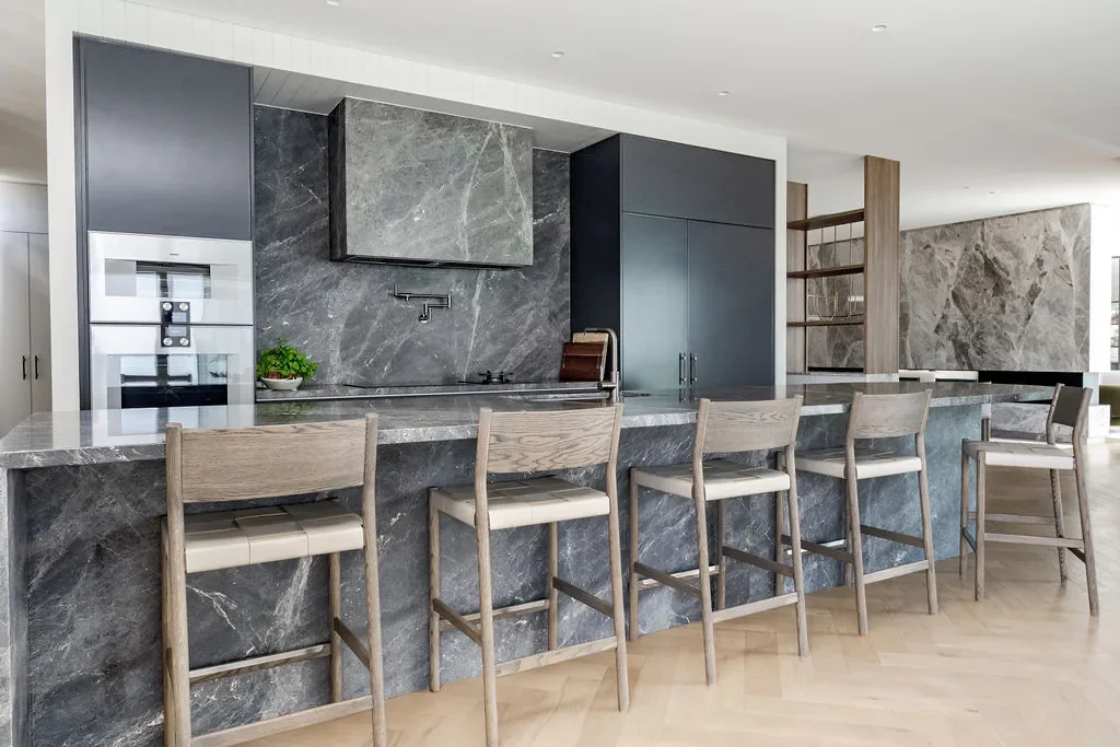 Modern kitchen with gray marble countertops and backsplash, gray cabinets, five wooden bar stools with white seats, built-in oven, and open shelving.