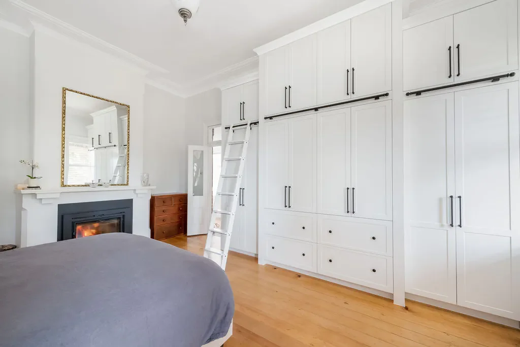 A bedroom with a bed covered in a gray blanket, a fireplace with a mirror above it, a wooden dresser, and large white built-in cabinets with black handles. A ladder leans against the cabinetry, and there is a small window near the ceiling.