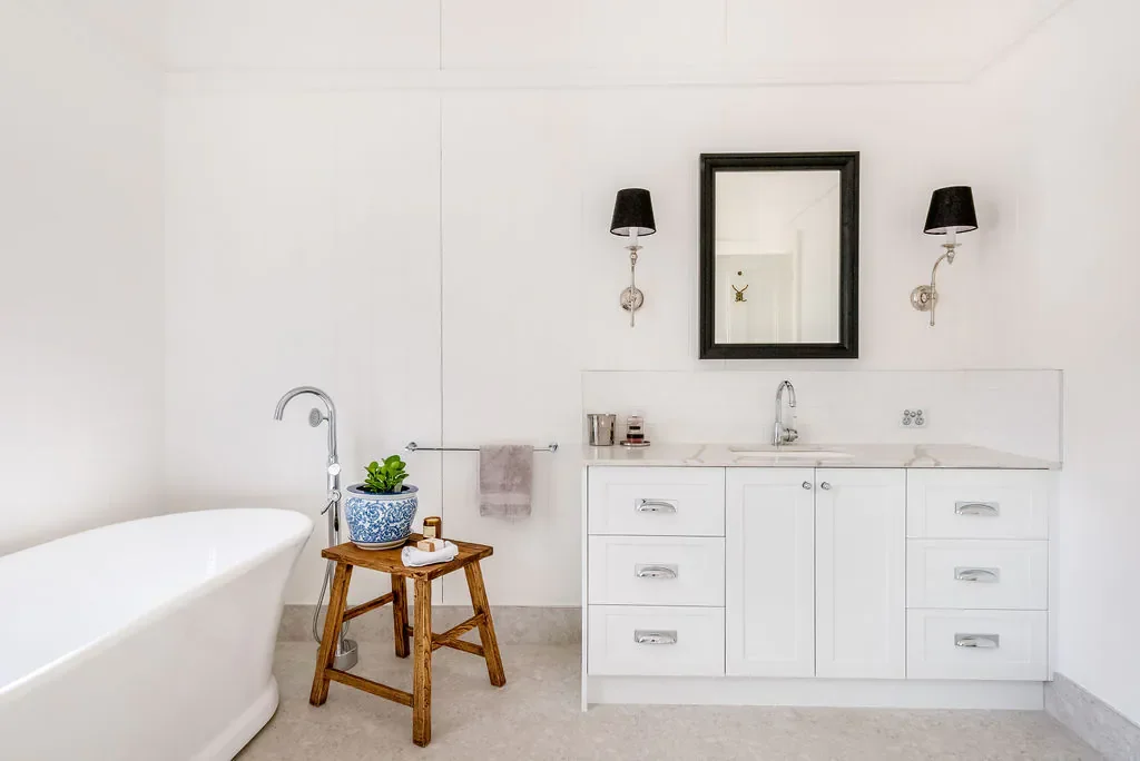A bright, minimalist bathroom with a white bathtub on the left, a small wooden stool with a potted plant and toiletries, a towel bar with a towel, and a white vanity with a sink. There is a mirror flanked by two wall sconces with black lampshades.