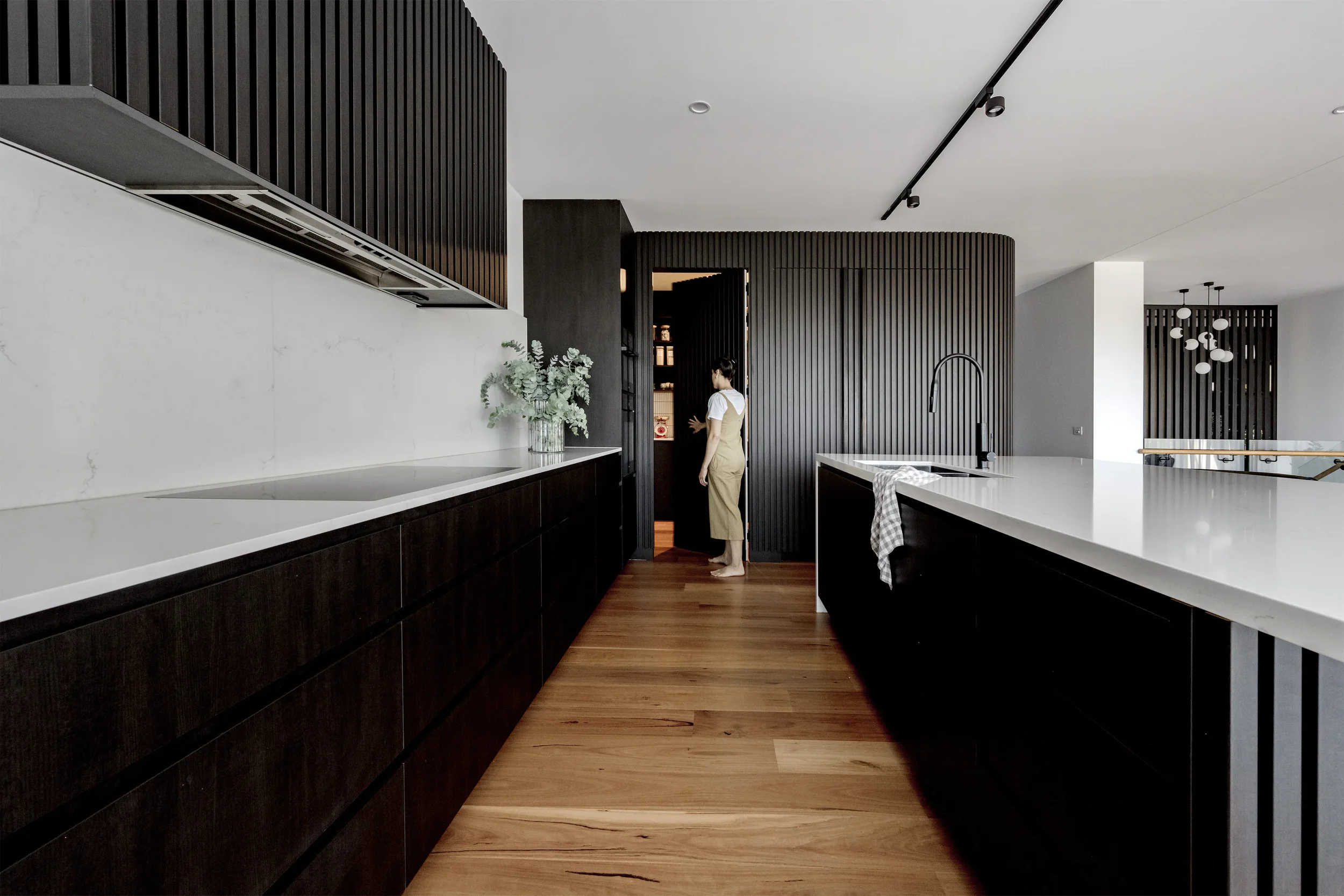 Modern kitchen with dark wood cabinets, white marble countertops, and a woman standing near the doorway. There are wooden dining floors, a vase with flowers on the counter, and a unique light fixture with multiple globes hanging from the ceiling in the background.