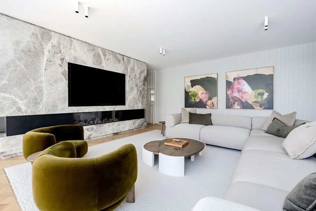 Modern living room with a large white sectional sofa, two abstract paintings on the wall, a marble accent wall with a mounted flat-screen TV, green velvet chairs, a round coffee table, and a white rug.