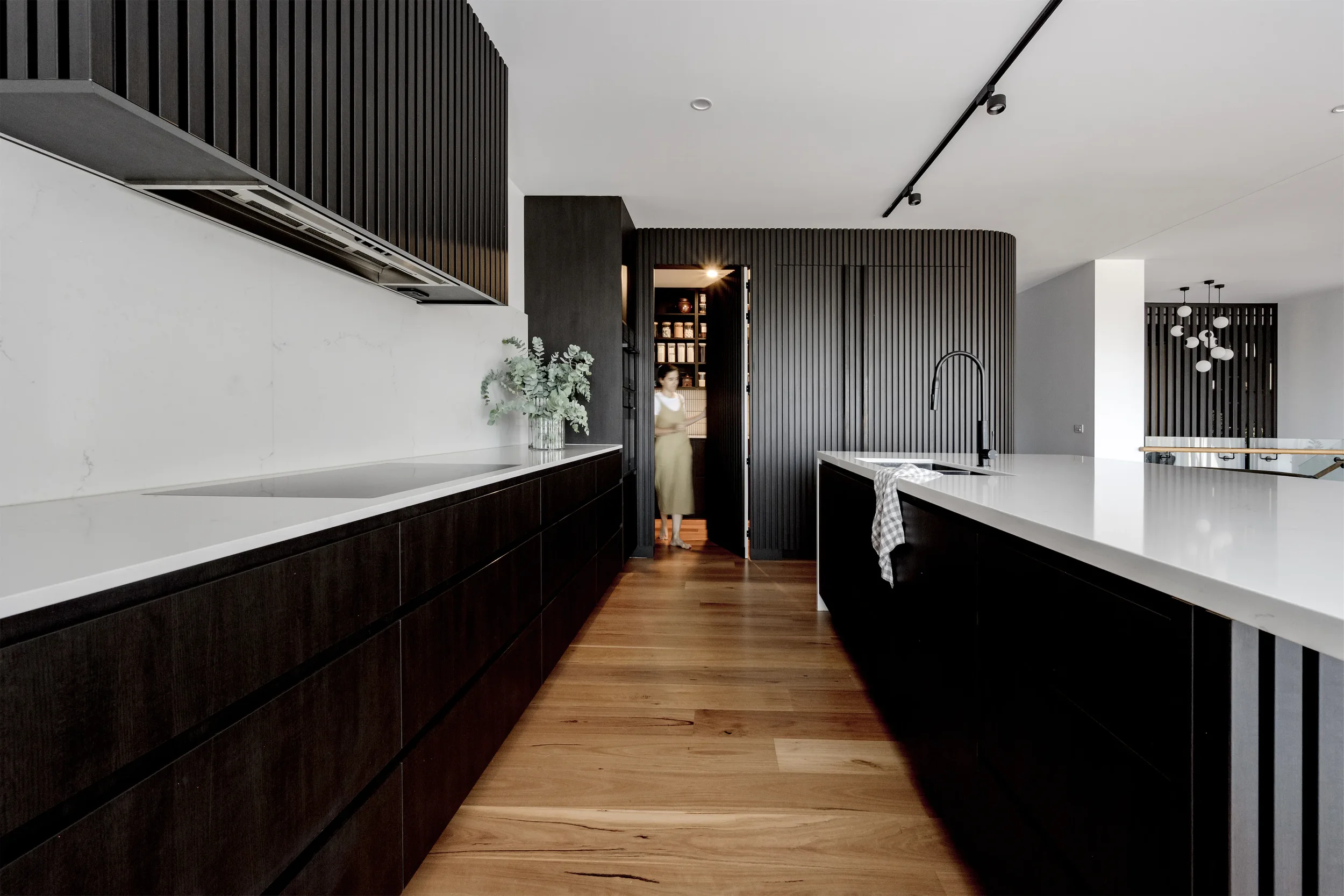 Modern kitchen with black cabinetry, white countertops, a white wall with a plant, and a person standing in the doorway.