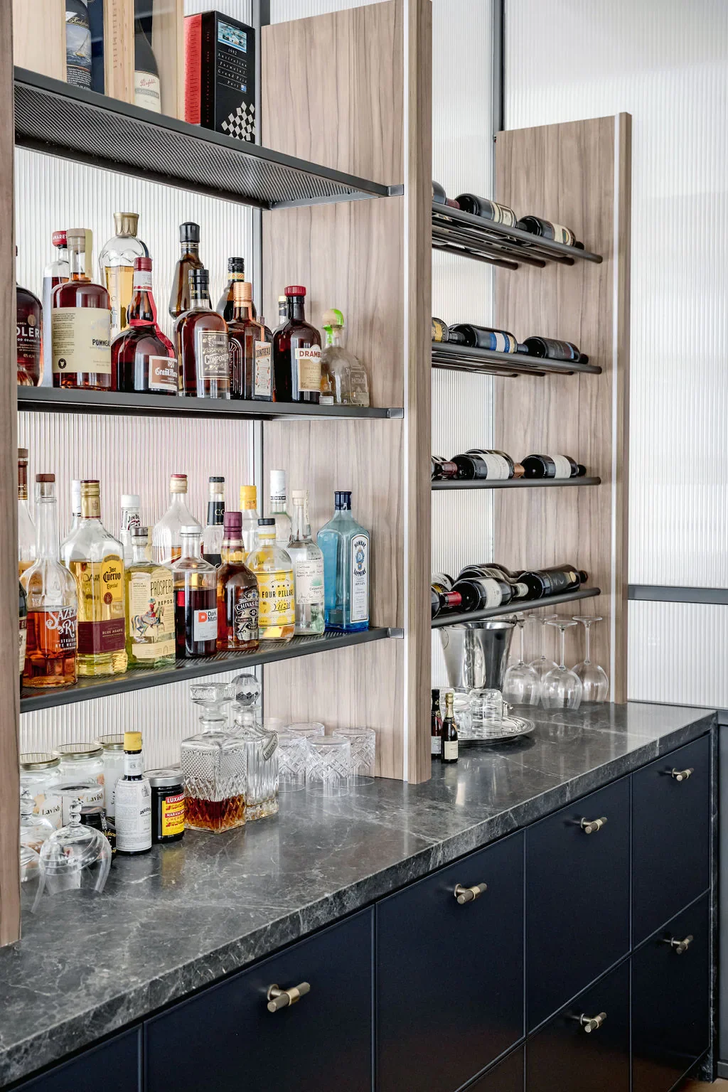 A bar with bottles of liquor on open shelves and a countertop with glassware and a wine cooler, against a light-colored wall.