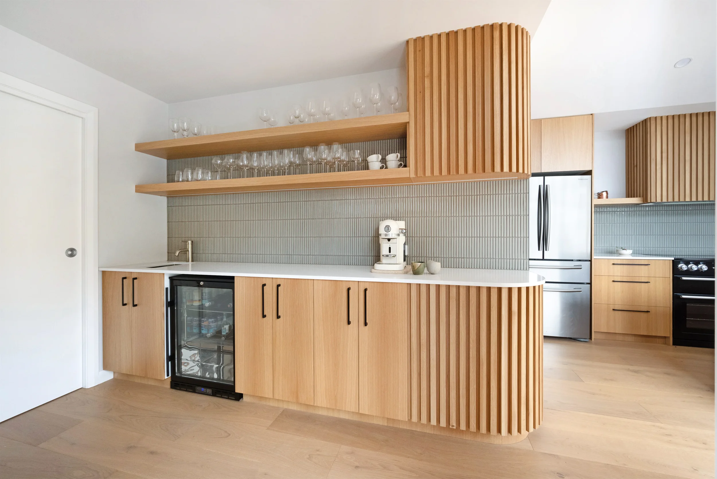 Modern kitchen featuring wooden cabinetry with vertical slats, open floating shelves with glassware and cups, a small wine fridge, a white countertop, and a stainless steel refrigerator, with light-colored wood flooring and minimal decor.