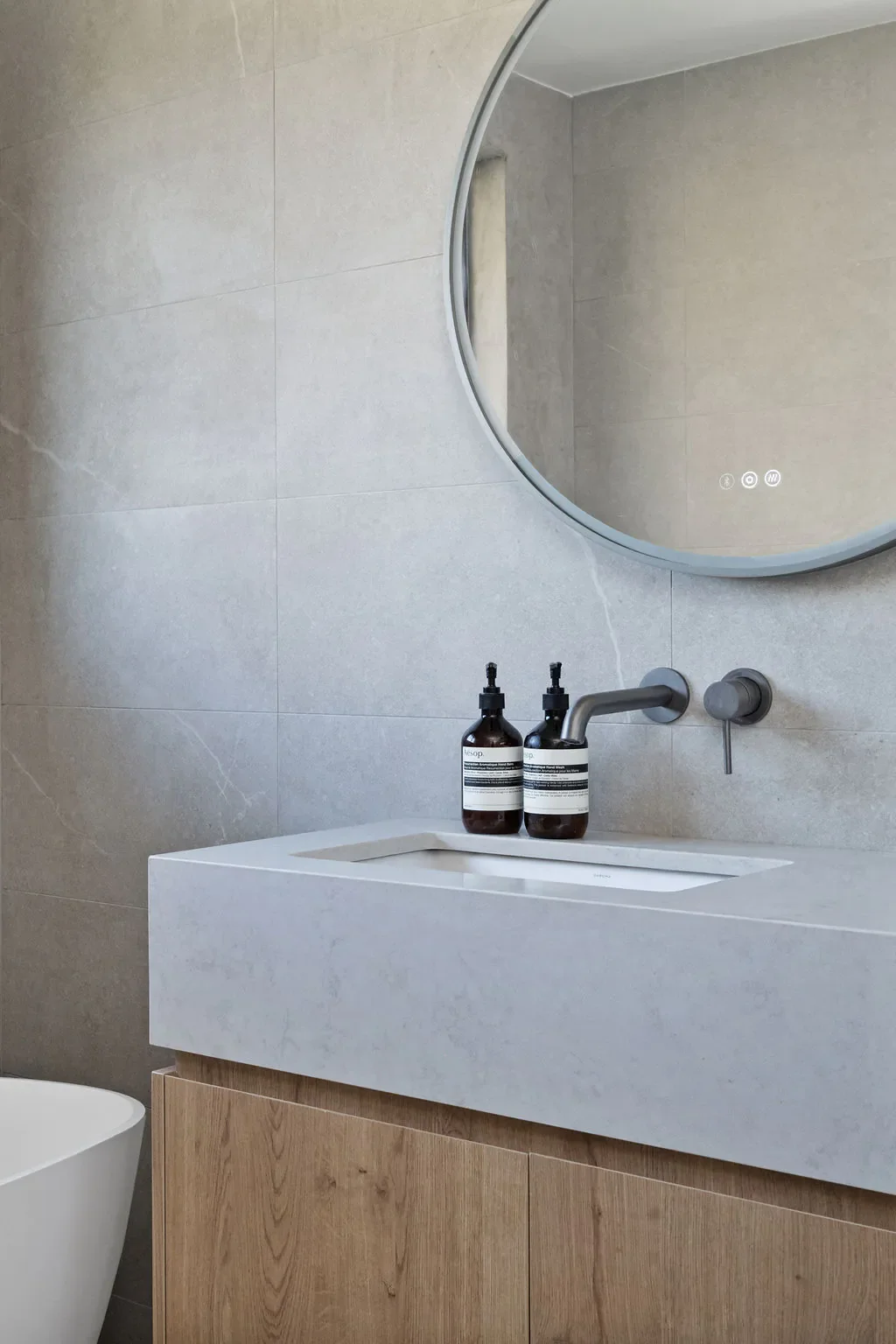 Bathroom mirror above a white sink with a wooden cabinet, two black soap bottles with pumps, gray walls, and a round mirror.