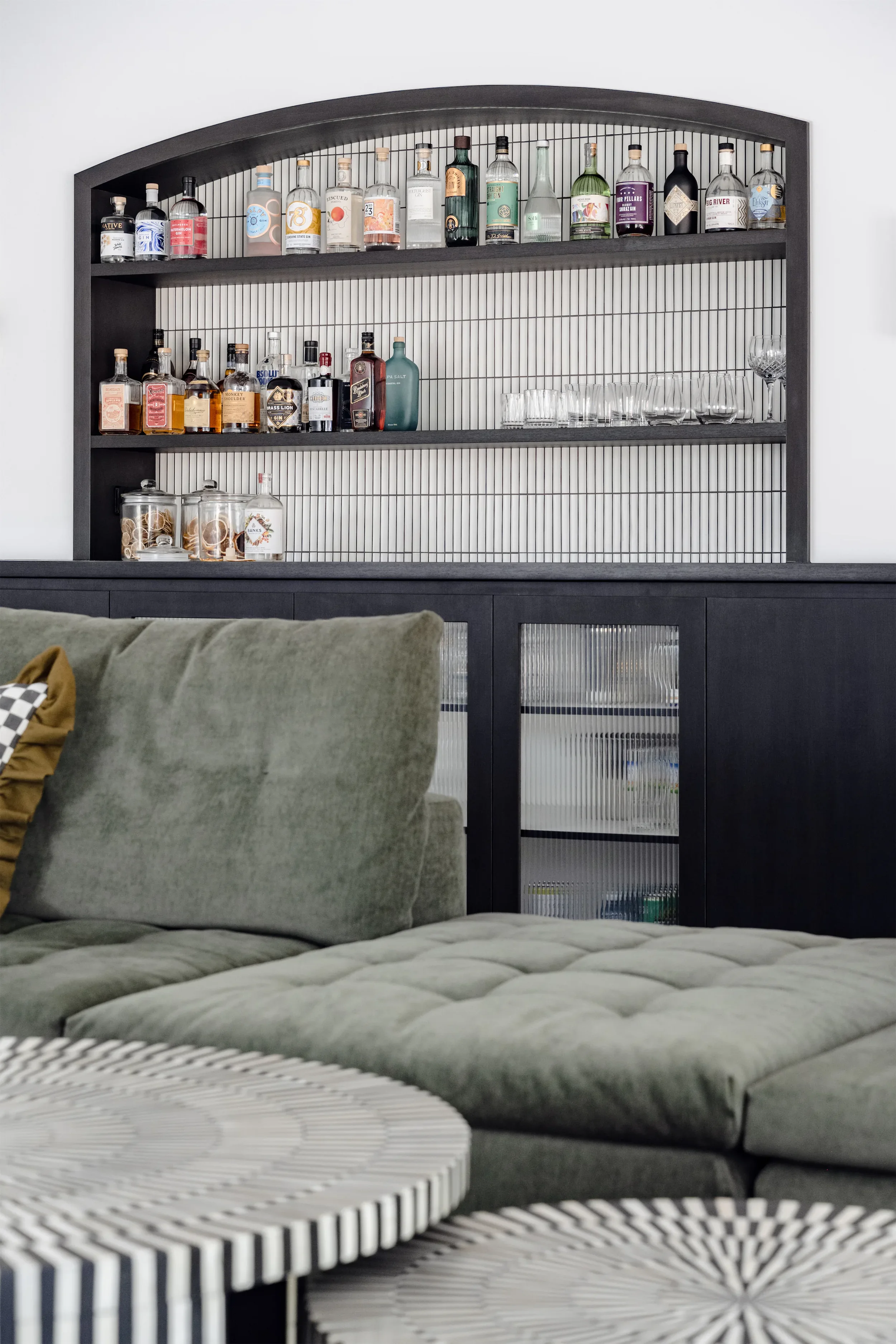 A home bar with shelves of liquor bottles and glassware, a green sofa with pillows, and a striped coffee table.