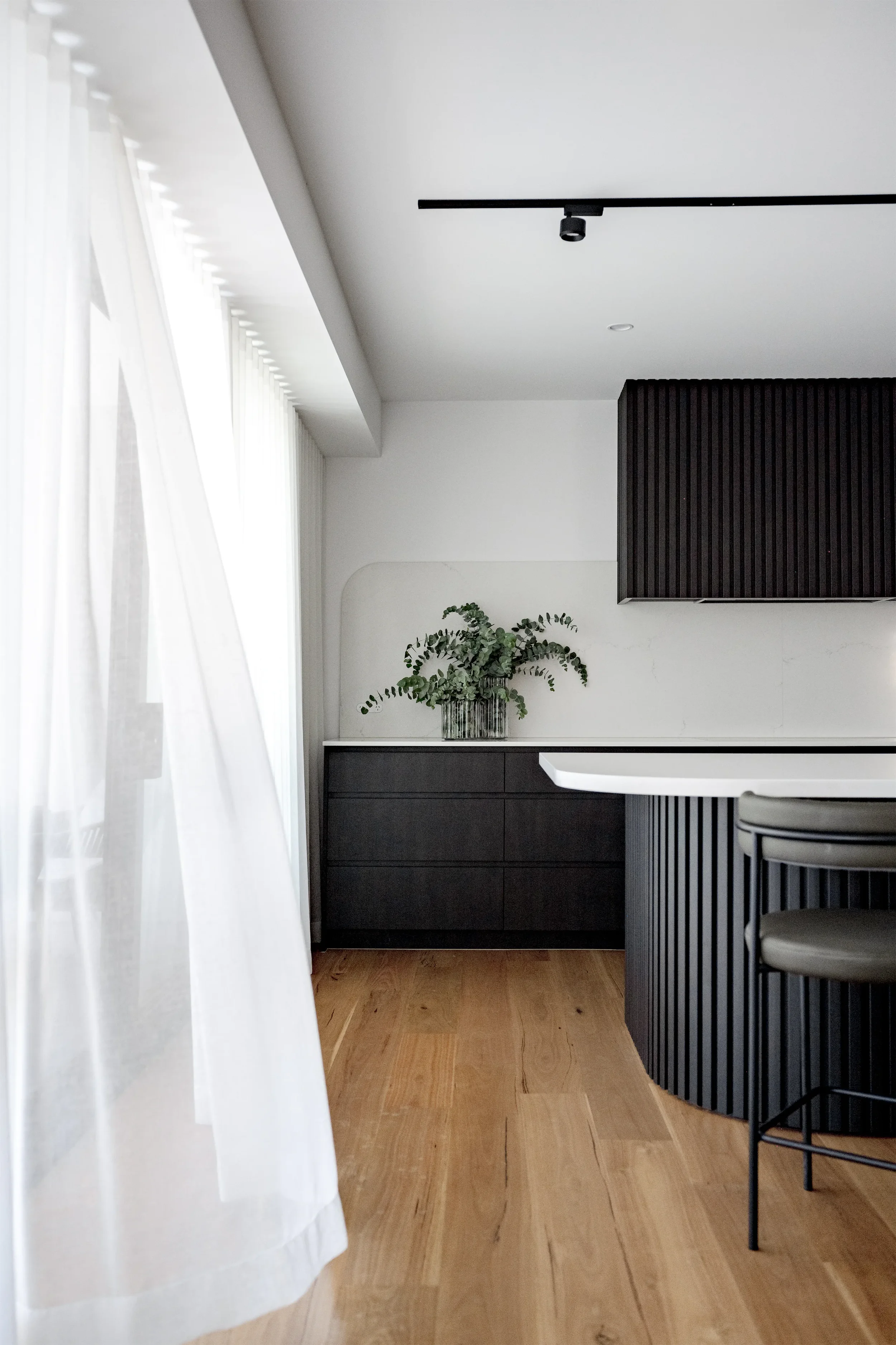 Interior view of a modern, minimalist room with white curtains on a window, a black cabinet or sideboard, a potted green plant, a hanging black track light, and black vertical slatted paneling on the wall. Wooden flooring.