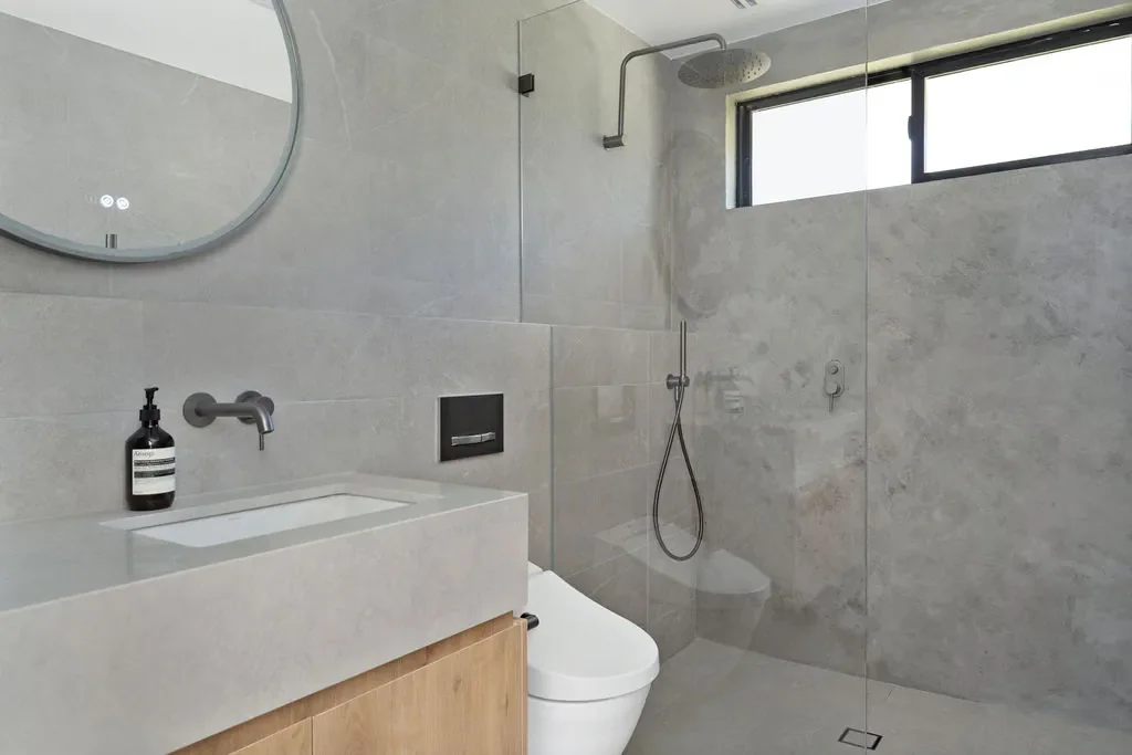 Modern bathroom with a walk-in shower, a wall-mounted toilet, a sink with a dark soap dispenser, an oval mirror, and a narrow horizontal window.