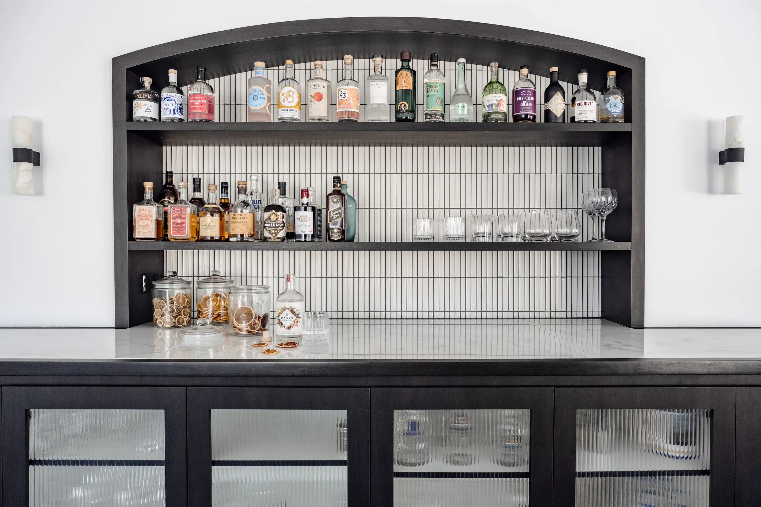 A black open-shelving bar unit with bottles of alcohol, glasses, and jarred garnishes on a white countertop with a white wall behind.