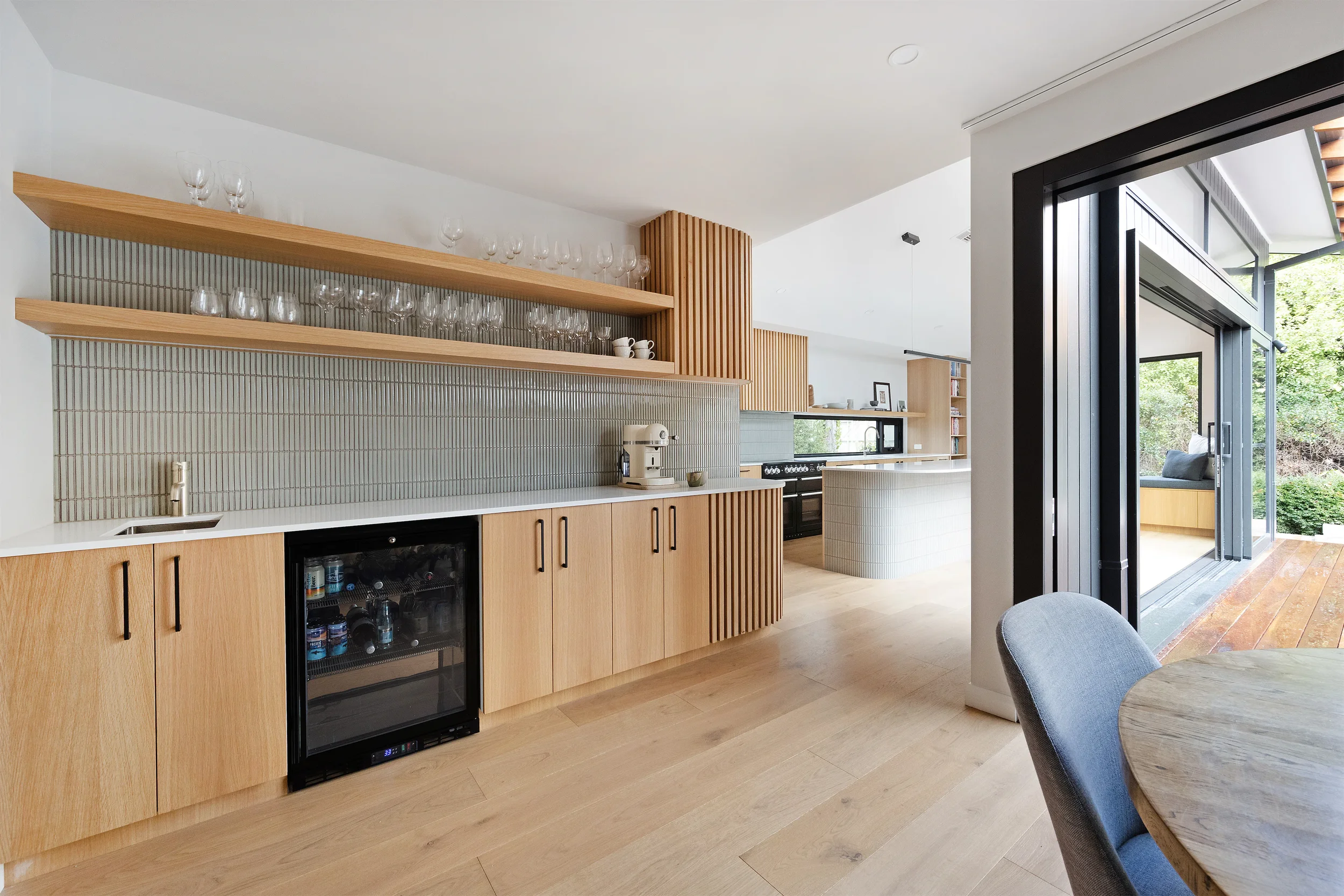 Modern kitchen with wooden cabinetry, open shelves with glassware and cups, a wine cooler, and sliding glass doors leading to an outdoor sitting area with greenery.
