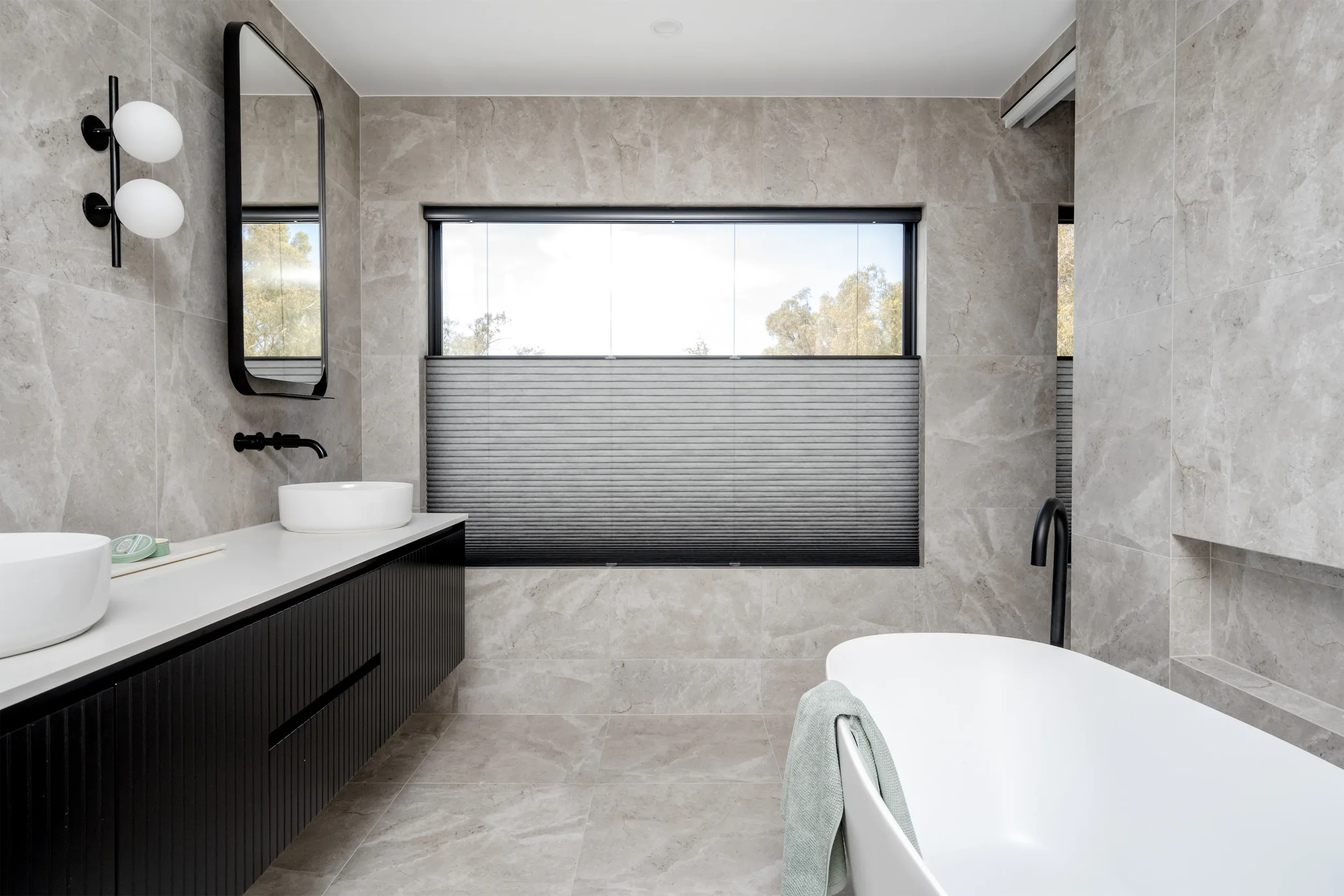 Modern bathroom with large beige tiles, a black vanity with two white vessel sinks, round wall-mounted light fixtures, a rectangular mirror, a large window with a roller shade, and a bathtub with a black faucet.