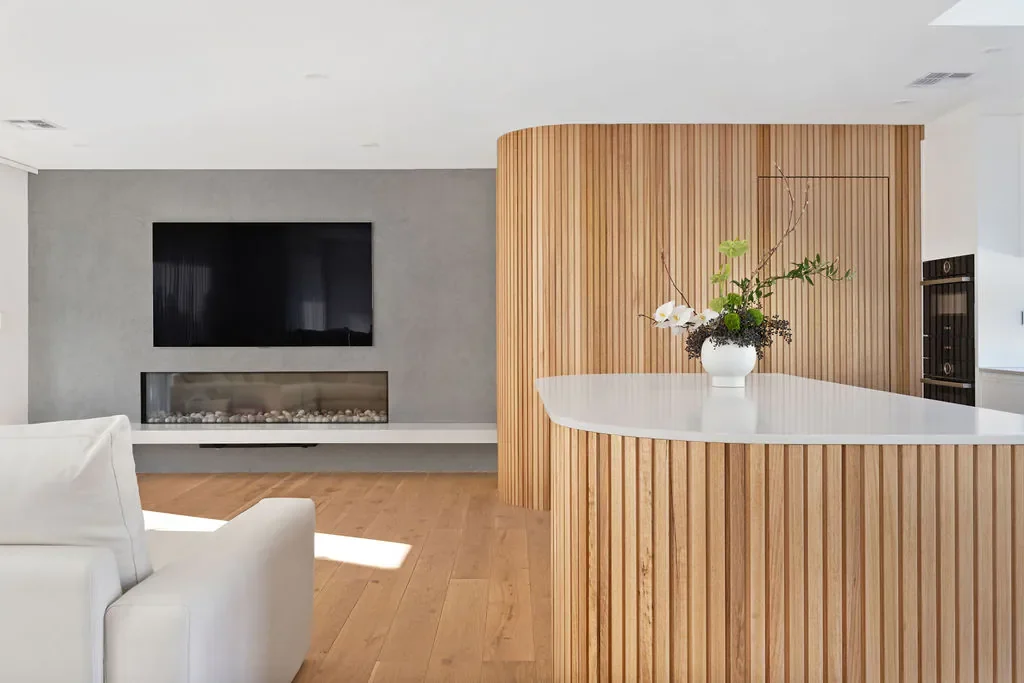 Modern living room with a white couch, a wall-mounted flat-screen TV, a fireplace, and a curved wooden divider with a flower arrangement on a white countertop.