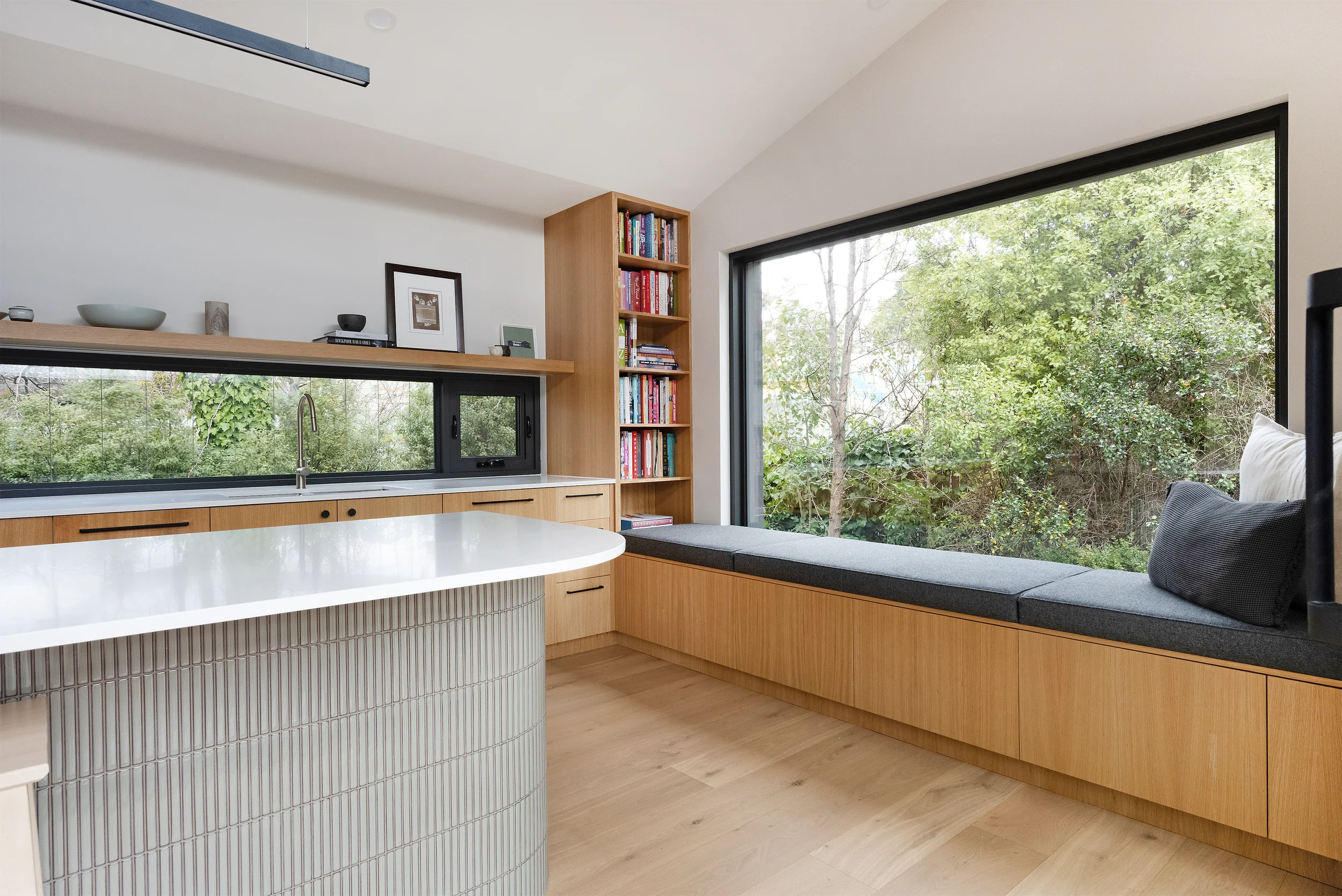 Modern kitchen with large window view of green trees, wooden cabinets, bookshelves, and a white countertop.