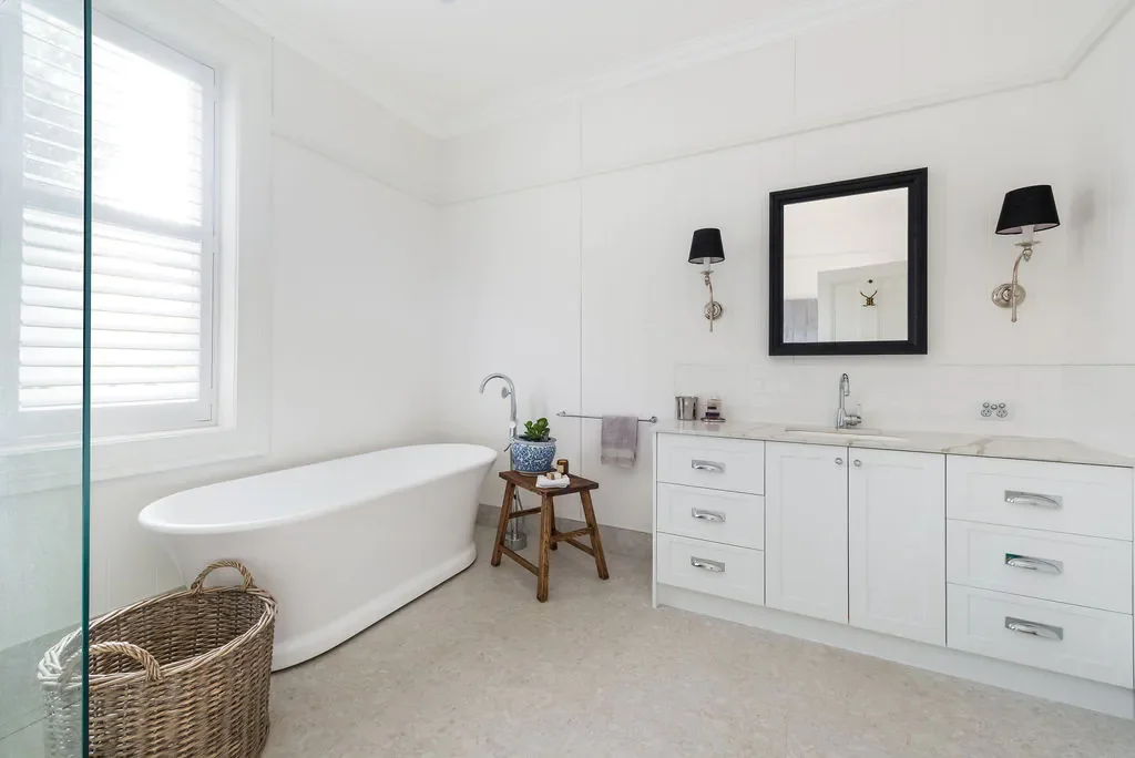 A modern bathroom with a white bathtub, window with blinds, wicker laundry basket, white vanity with drawers, a mirror, wall sconces, and a small wooden stool with a plant.