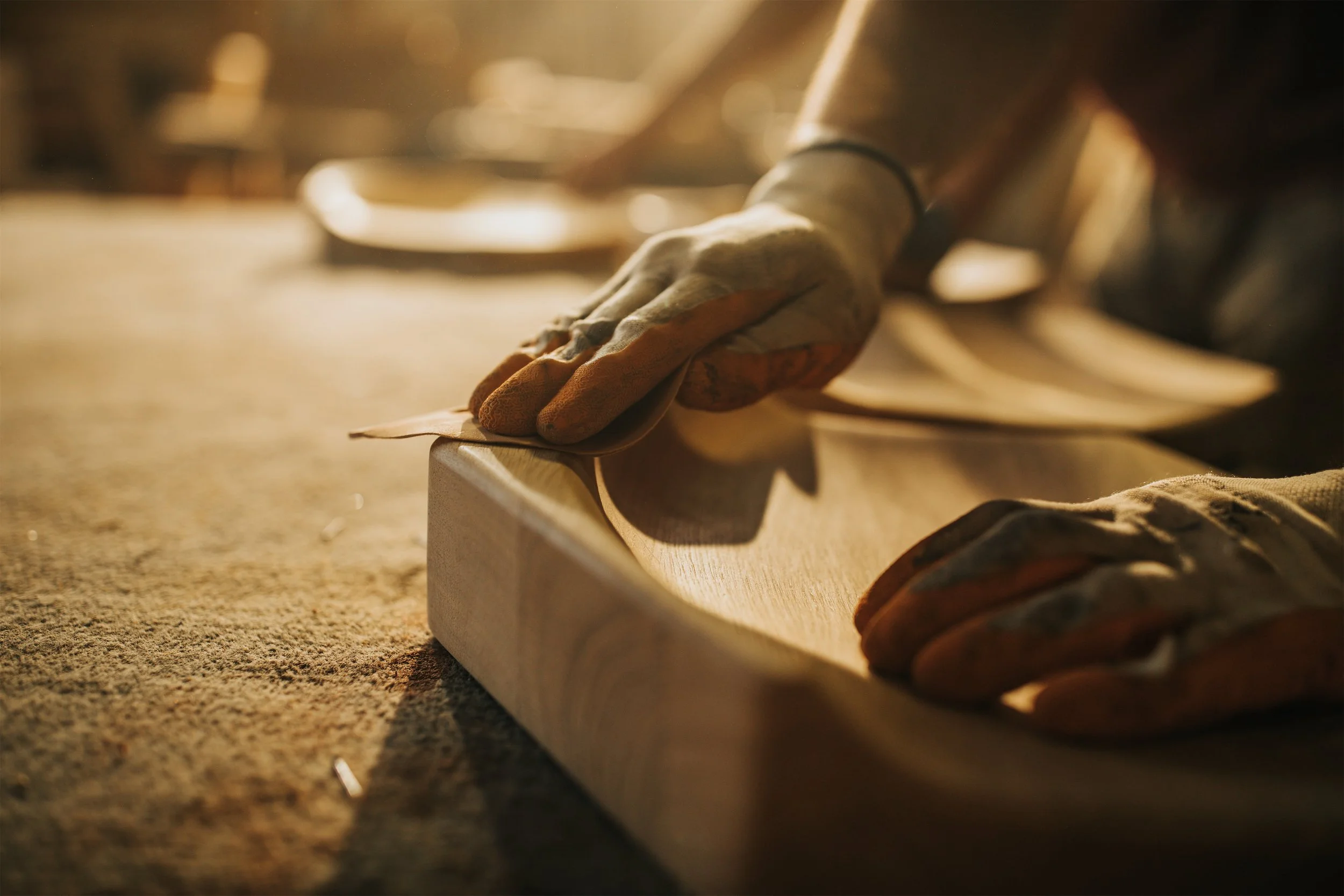Close-up of a hand wearing a glove, working on a piece of wood with a tool in a warm, well-lit workshop.