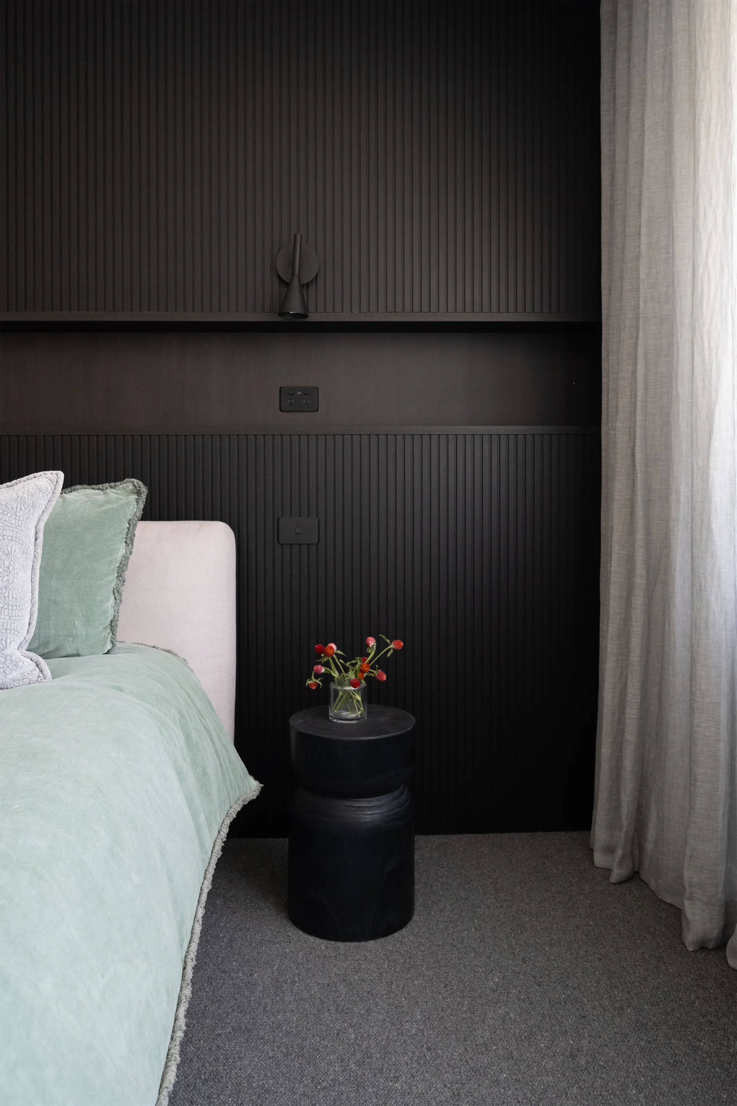 A bedroom corner with a bed featuring light green and white pillows, a black bedside table with a glass vase of red and pink flowers, a dark paneled wall with a wall-mounted lamp, electrical outlets, and a window with light-colored curtains.