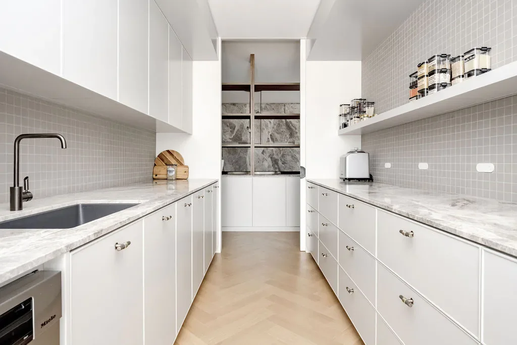 Modern kitchen with white cabinets, marble countertops, gray tile backsplash, and wooden floor. Shelves hold spice jars, small appliances are on the right counter, and a small sink is on the left.