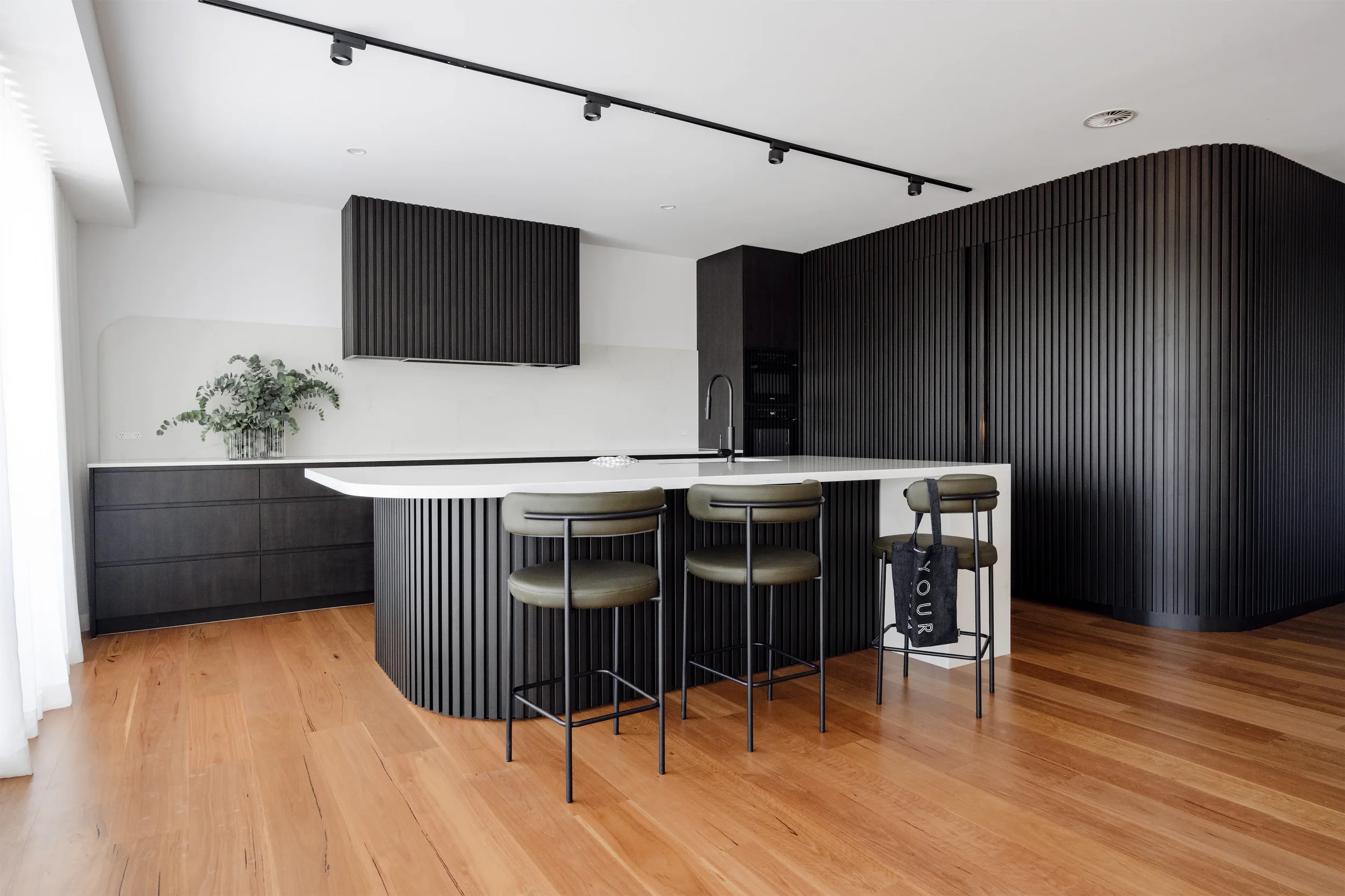 Modern kitchen with black vertical slats, white countertops, gold barstools, a potted plant on the counter, and a hardwood floor.