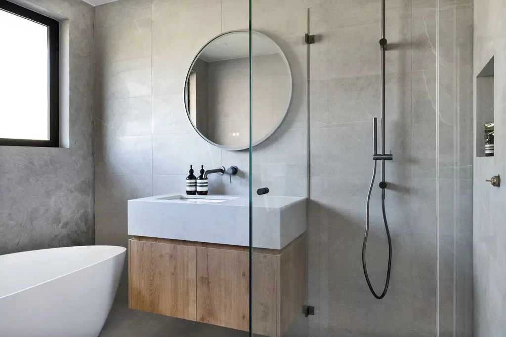 Modern bathroom with a bathtub, a white sink with wooden cabinet, a large round mirror, a window, and a walk-in shower with a glass partition.