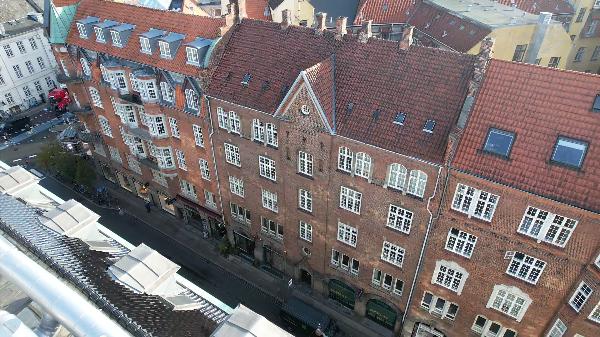 Et højt synsvinkelbillede af en bybygning med røde murstensfacader og tag med tegltagsten. Gaten nedenfor har cykler og biler, og der er butikker i stueetagen af bygningen.
