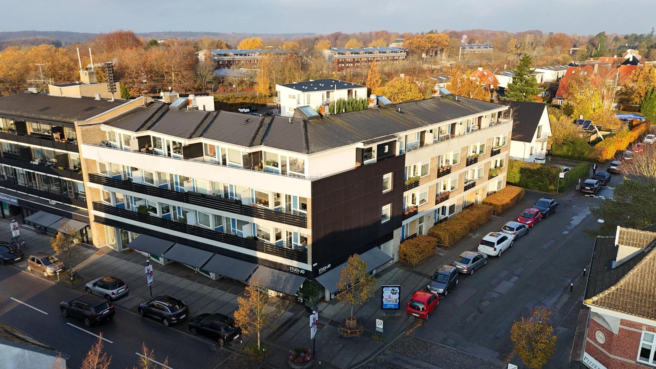 Et luftfoto af en by med flere boligblokkere, parkeringspladser og veje, omgivet af træer med efterårsfarver.