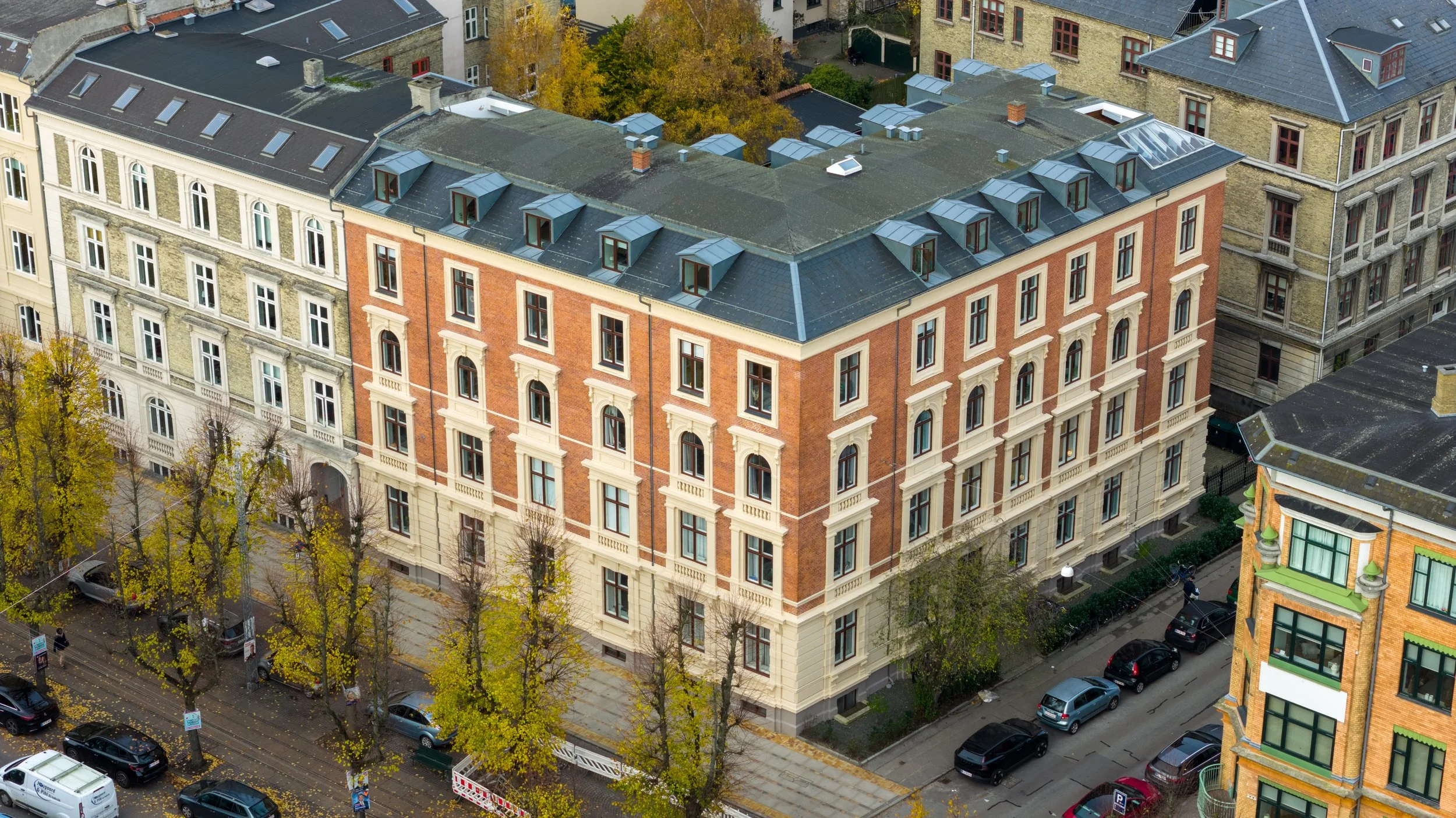 Et højt bygning med rød murstensfacade, mange vinduer og tag med flere ovenlysvinduer, omgivet af træer med efterårsfarver og andre bygninger. Gade med parkerede biler foran.
