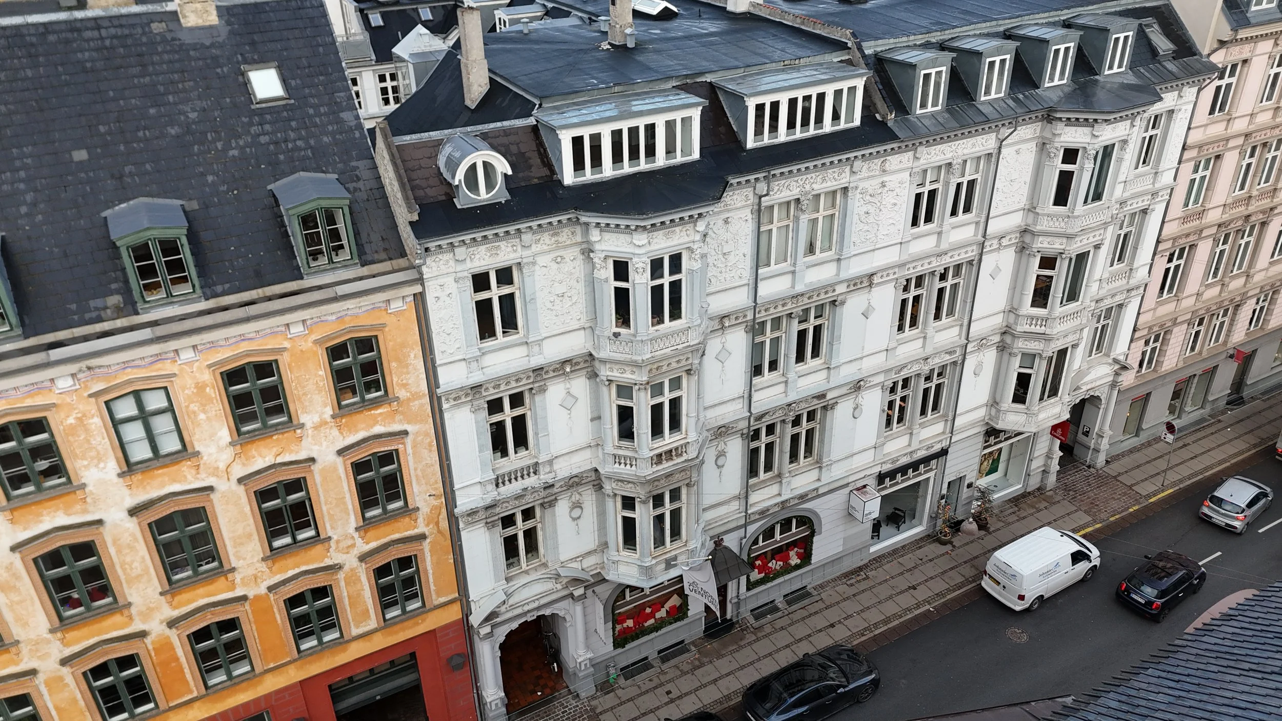 Udendørs bygningsfacade med flere historiske bygninger med store vinduer, detaljerede udskæringer og forskellige farver på facaderne, set fra oven.