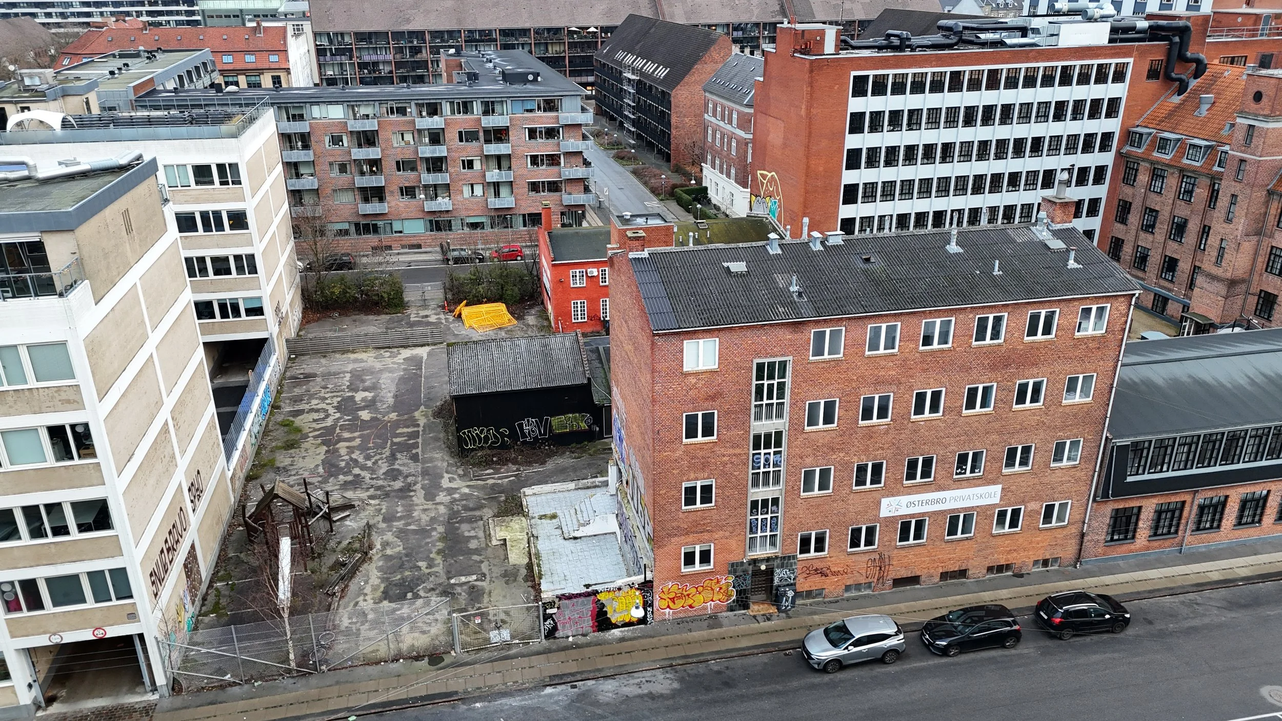 Et højhuskompleks med flere bygninger, herunder murstensbygninger med graffiti, en mur med træningsudstyr og biler parkeret langs vejen, set fra en høj højde.