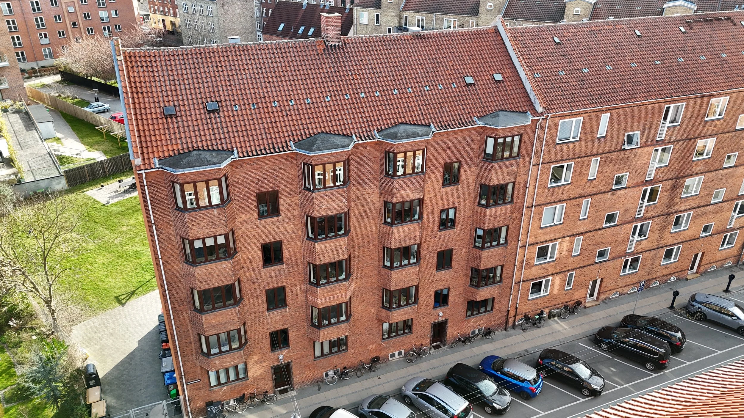Et højt, rødt murstenshus med flere rækker vinduer og et rødt tegltag, set fra oven. Der er parkerede biler og cykler foran bygningen, samt en grøn have og gangstier til venstre.