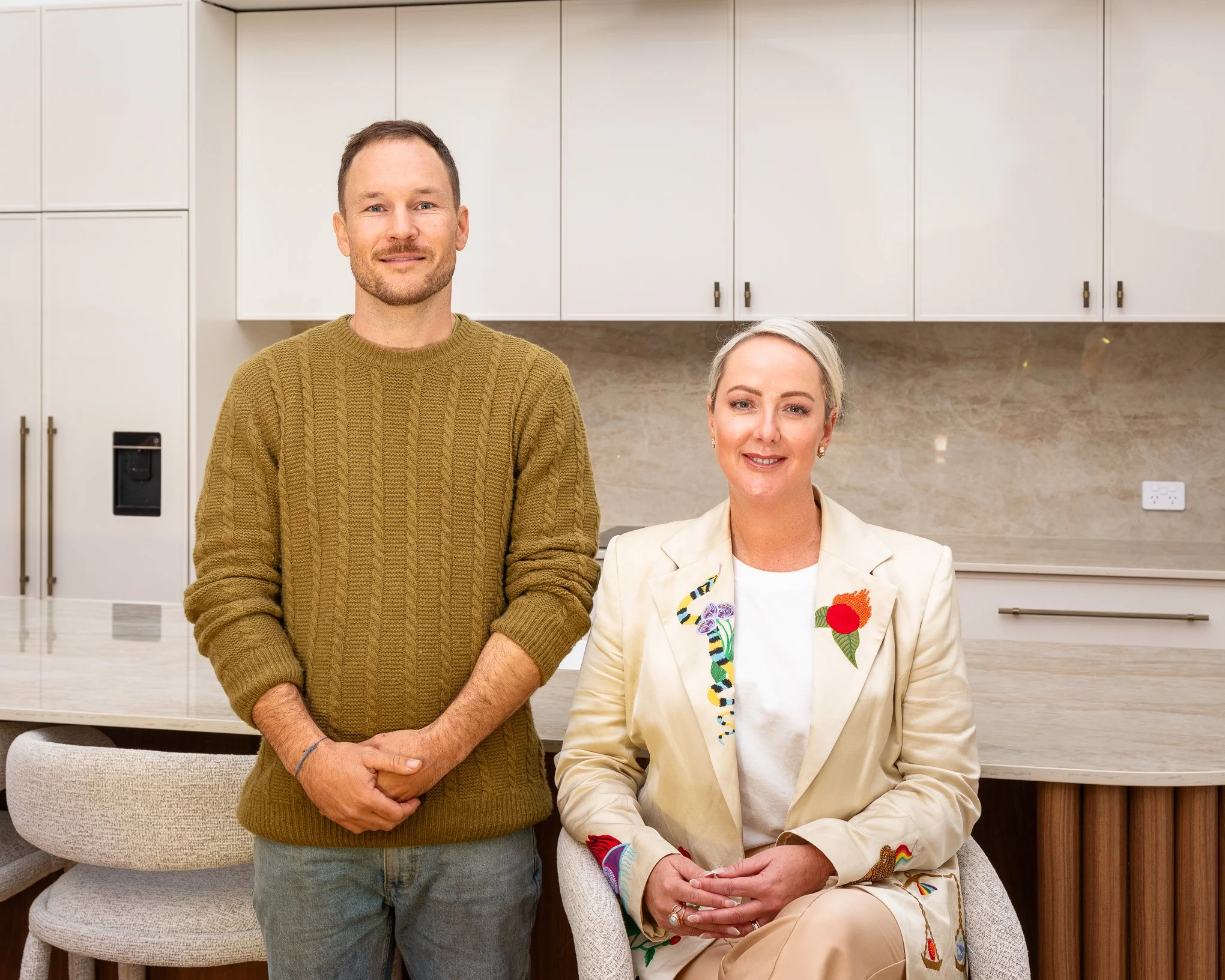 A man with light skin, short brown hair, and a beard wearing a brown knitted sweater, standing next to a woman with light skin and gray hair. The woman is seated and dressed in a beige blazer with various colorful embroidered patches, including a sna