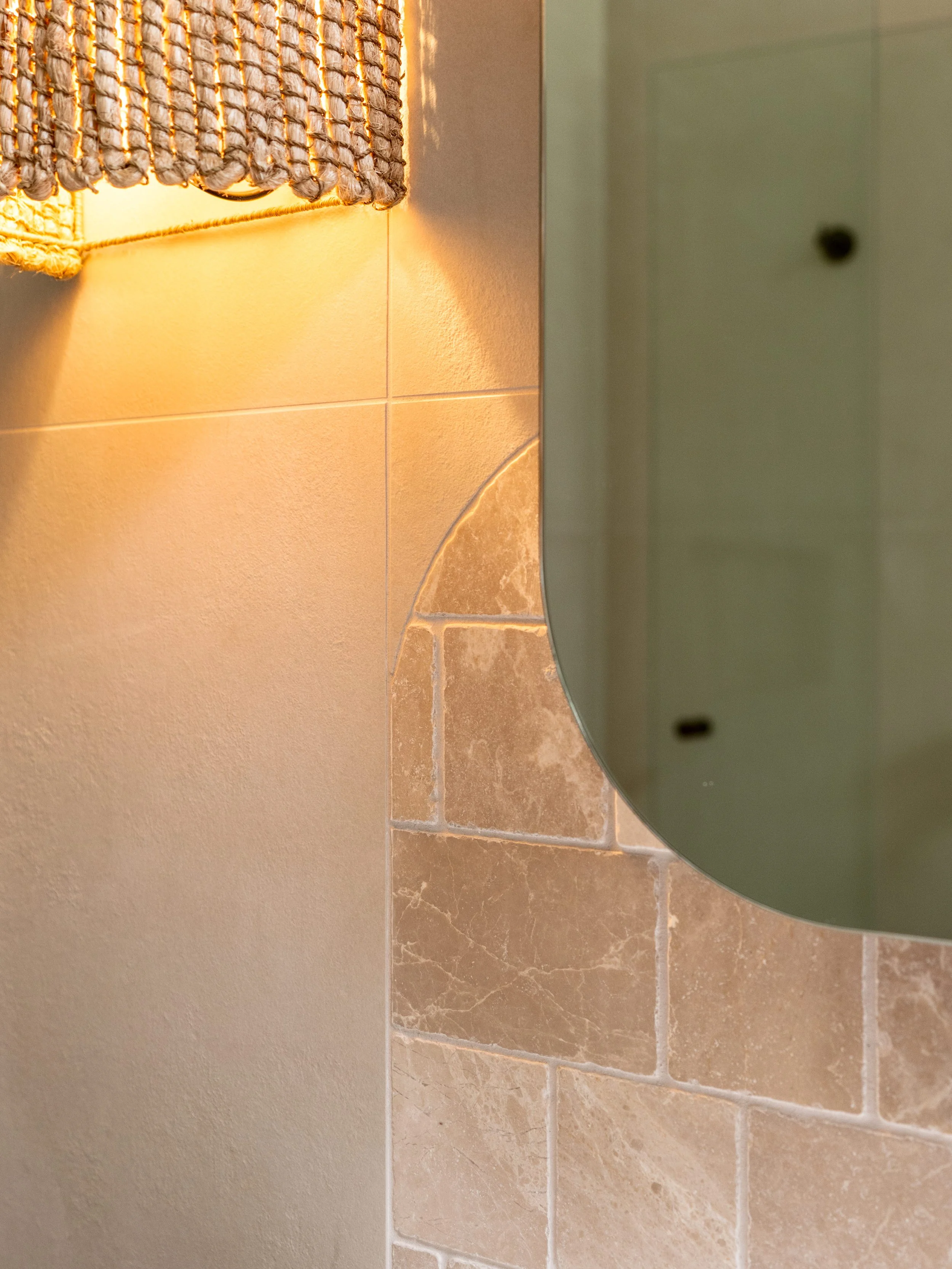 Close-up of a bathroom corner with a textured woven wall hanging, beige tiled wall, and a curved green mirror reflecting part of the area.
