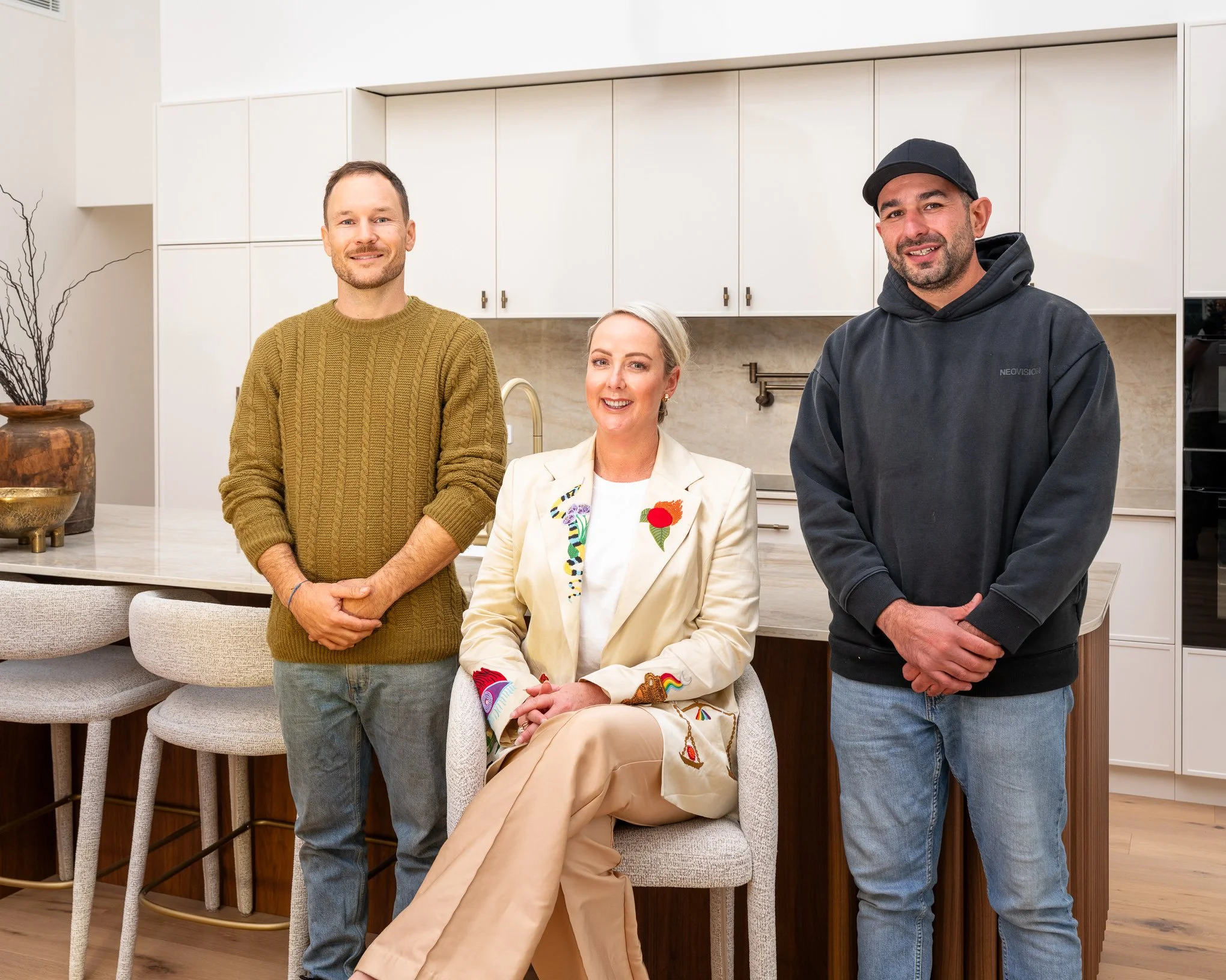 Three people standing in a kitchen with white cabinets and a beige backsplash, two men and one woman, all smiling and looking at the camera. The woman is sitting on a chair, dressed in a beige outfit with colorful embroidered details. The man on the 