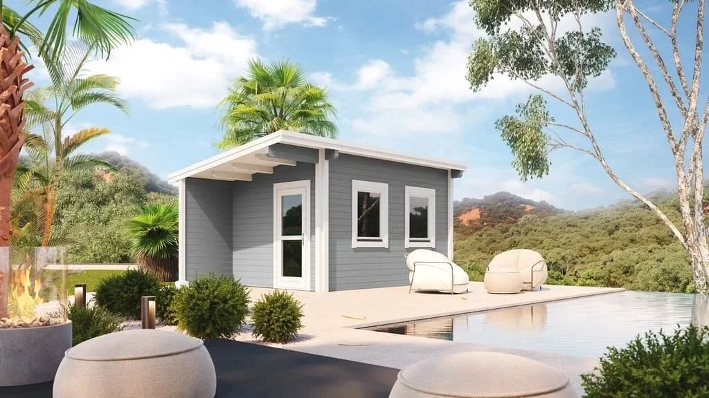 Small modern house with gray siding and white trim, surrounded by trees and plants, with an outdoor patio and seating area near a pool.