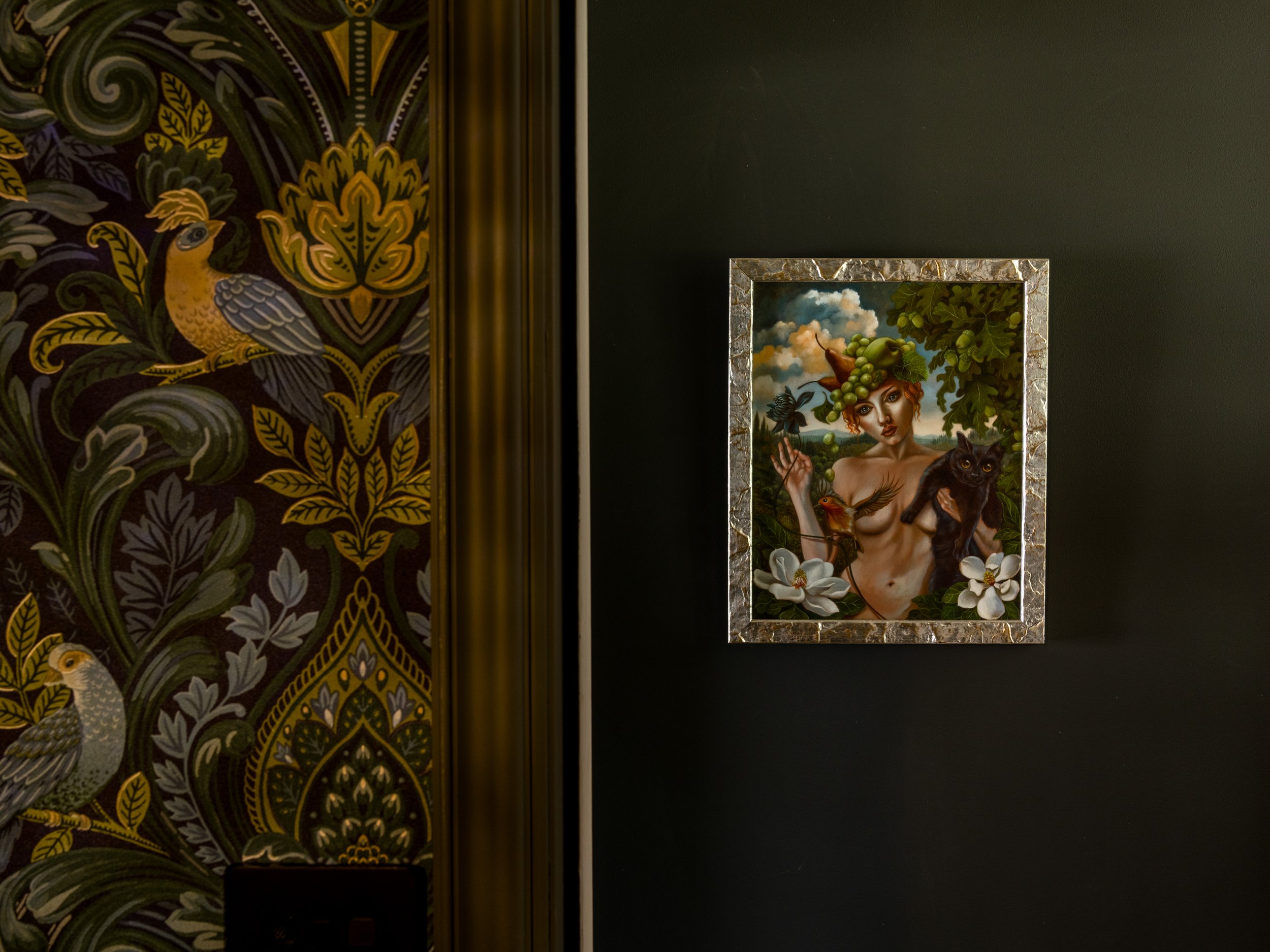 A framed painting of a woman with curly hair and a crown of green grapes, holding a small bird, with a black cat sitting beside her. The background depicts a sky with clouds and trees.