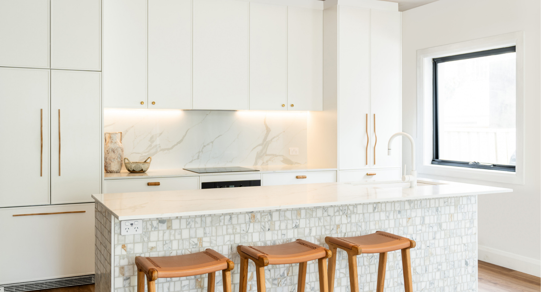 Modern white kitchen with marble backsplash and countertops, wooden barstools, and a large window with black frame.