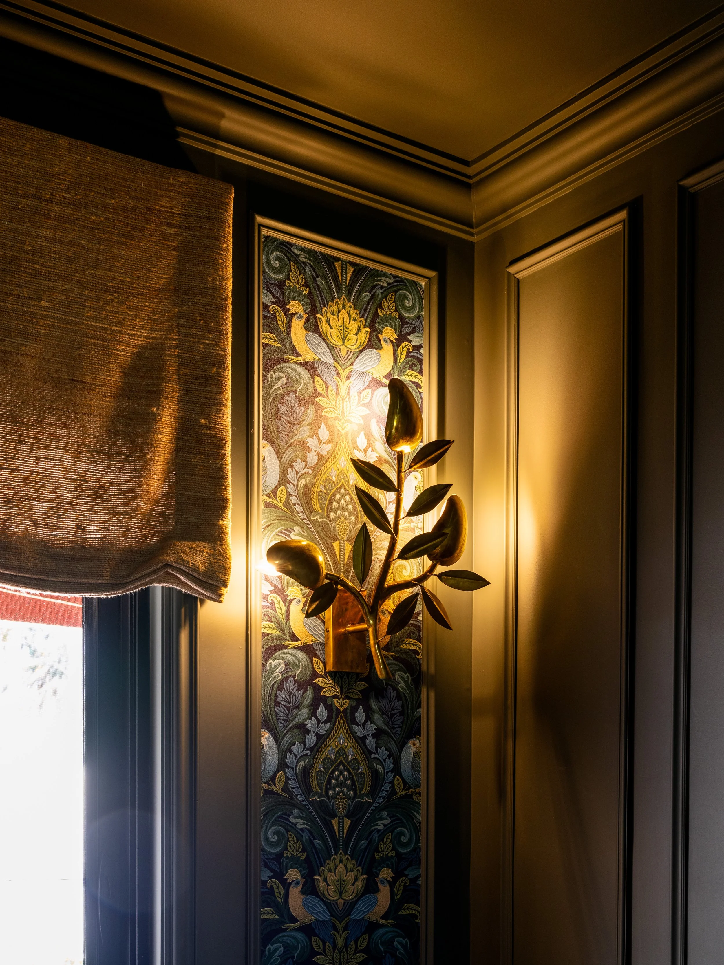 An interior corner with a dark wall panel, a decorative tapestry with peacocks and floral pattern, a metallic light fixture shaped like a leaf, and a window with a textured brown curtain.