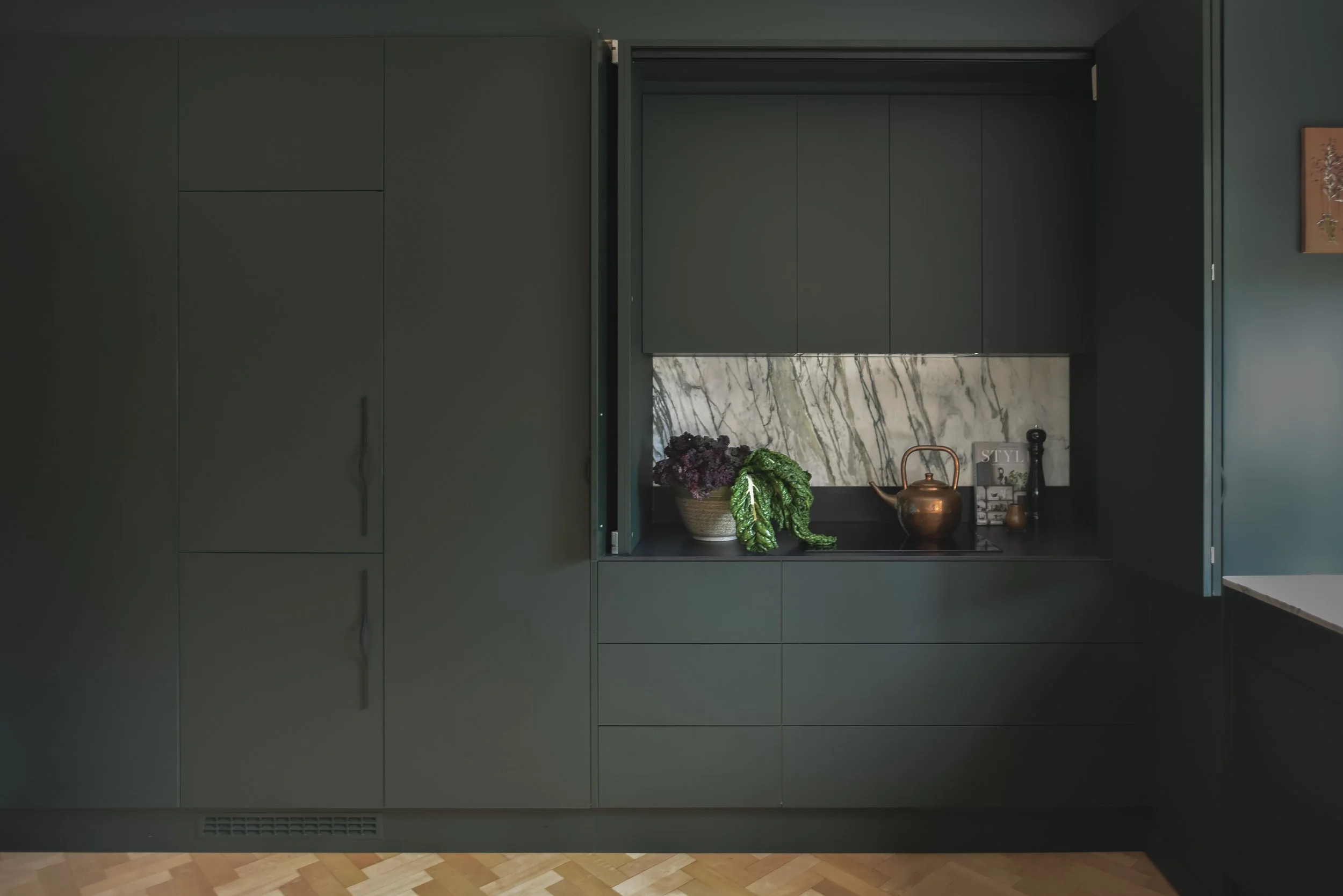 A section of a modern, dark green kitchen with closed cabinetry and a countertop featuring decorative items, including a basket of purple flowers, a green plant, a copper teapot, and a black bottle, against a marble backsplash.