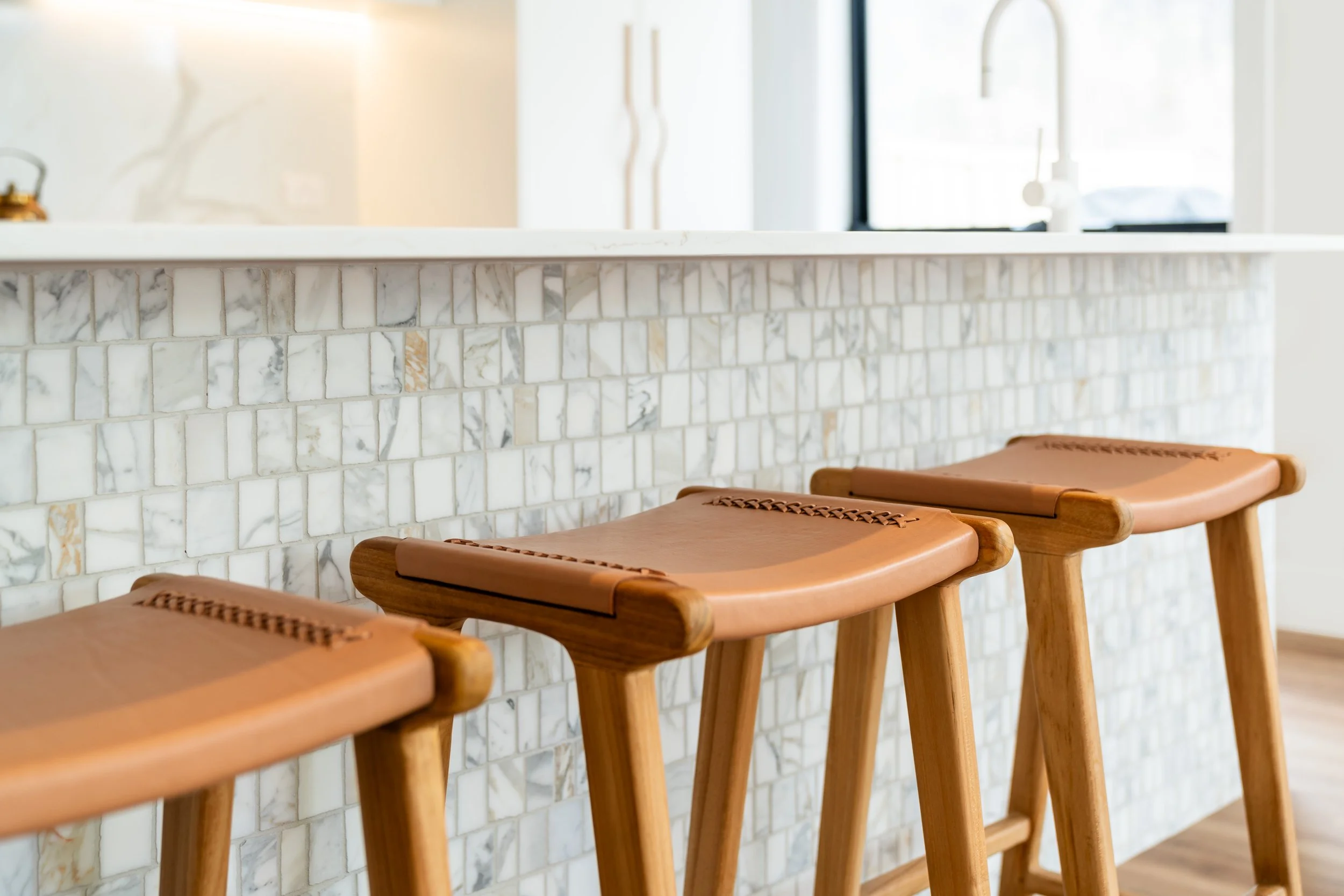 A row of four wooden bar stools with leather seats in front of a white marble kitchen island, with a white sink and faucet in the background in a modern kitchen with bright, natural lighting.