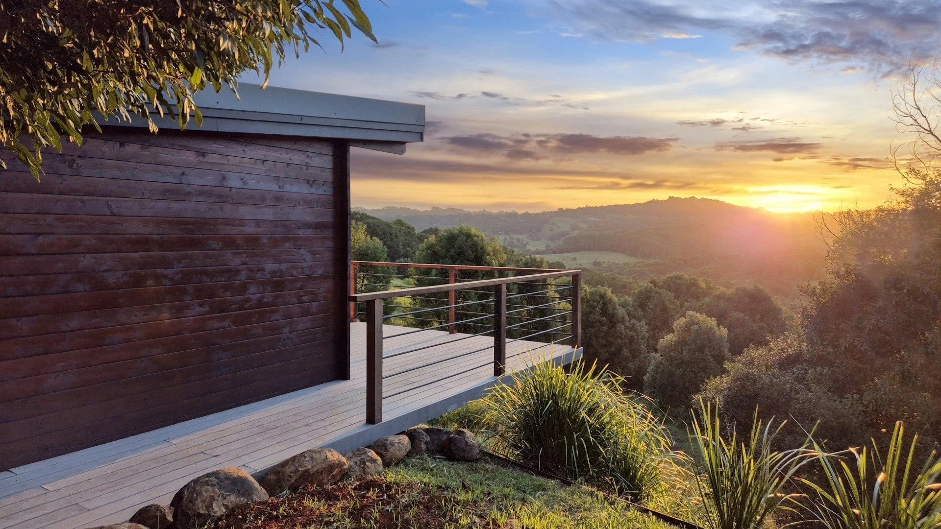 Sunset view from a balcony overlooking rolling hills, greenery, and a partly cloudy sky, with a wooden and metal railing and part of a wooden house or deck in the foreground.
