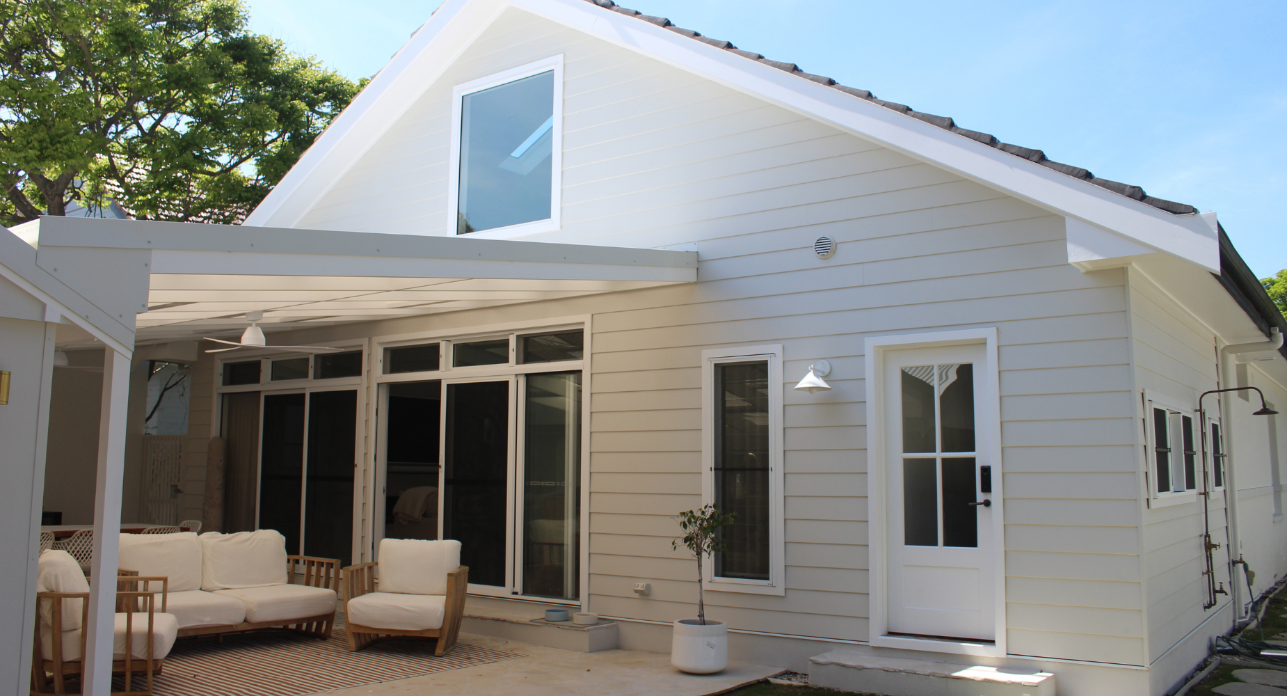 White house with dark roof and multiple windows, including a sliding glass door, in a backyard with outdoor furniture and a striped rug under a patio cover, a small potted tree, and a clear blue sky.
