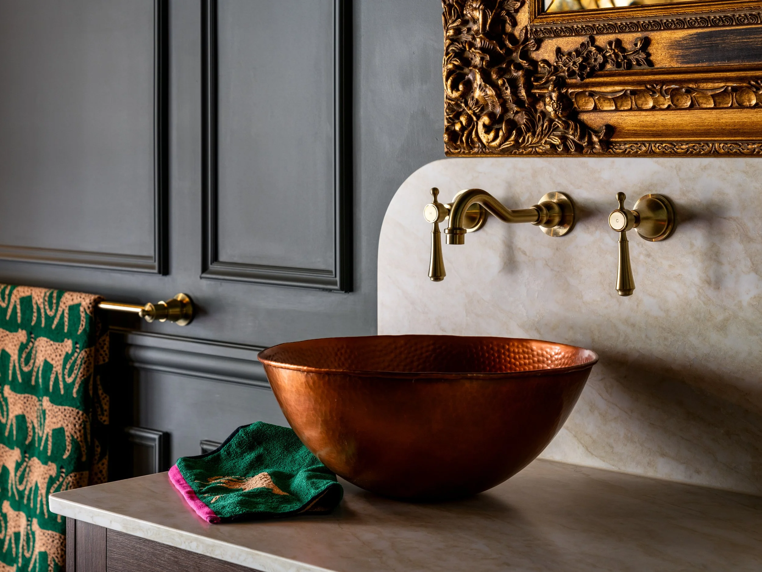 Decorative bathroom sink with a copper basin, gold faucet, marble countertop, and ornate gold framed mirror, with a green towel with pink trim on the counter.