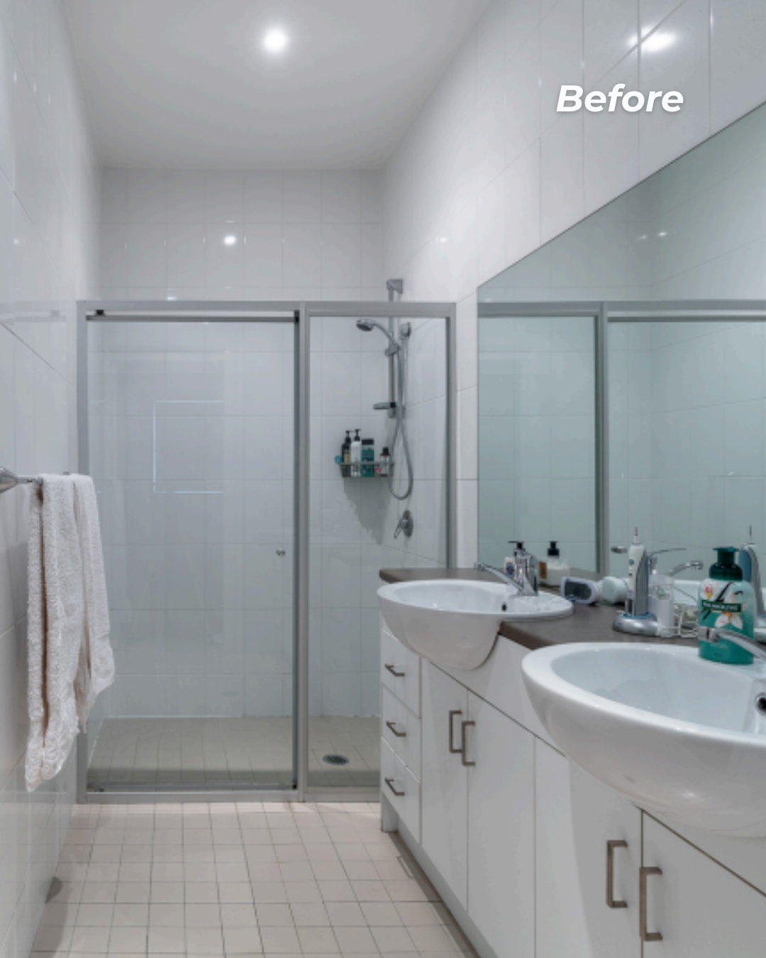 A bathroom with a glass shower enclosure, white tile walls, a double sink vanity with a mirror, and various toiletries on the counter.